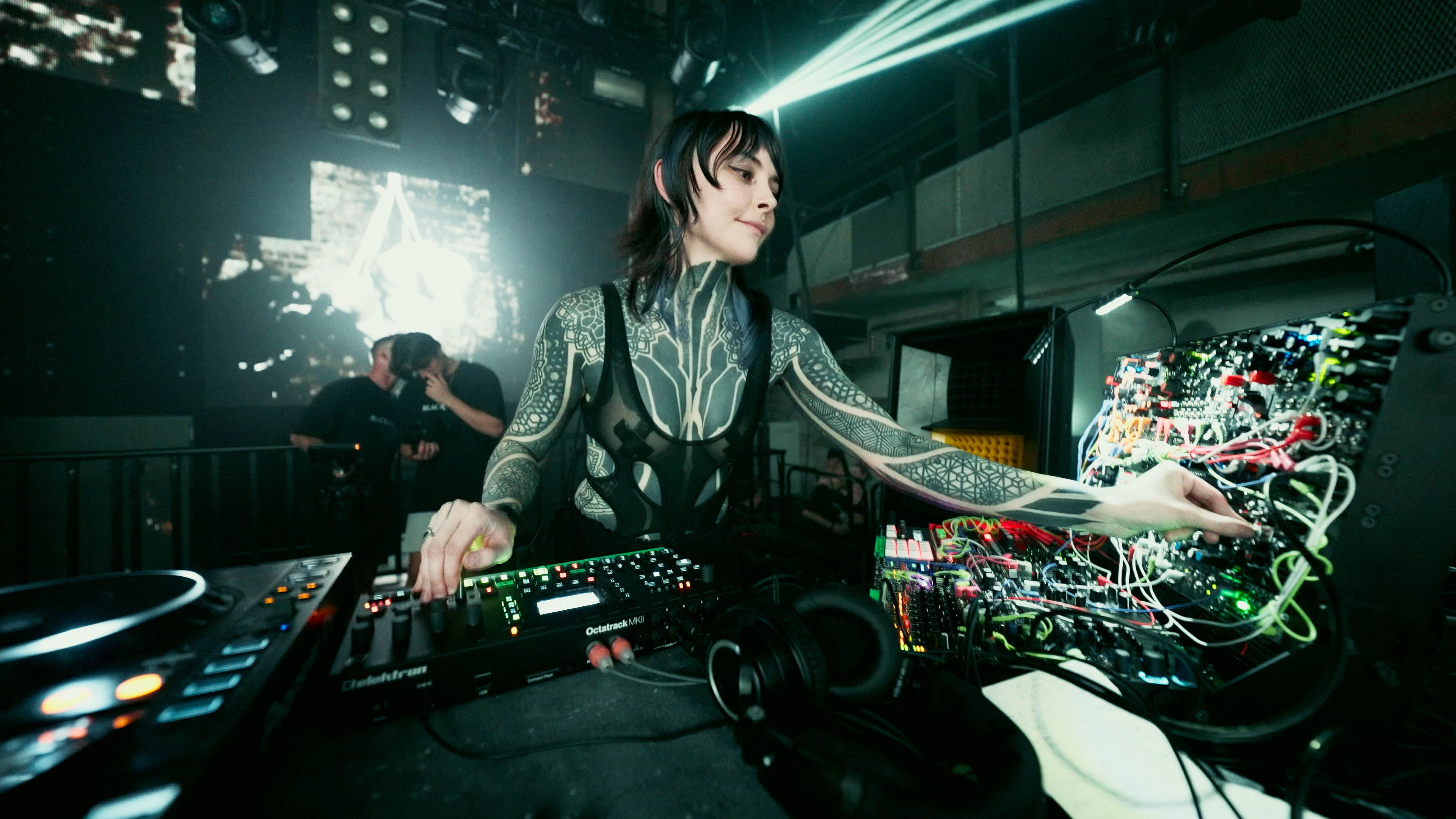 Krista performing with a modular synthesizer and DJ equipment beneath green stage lights