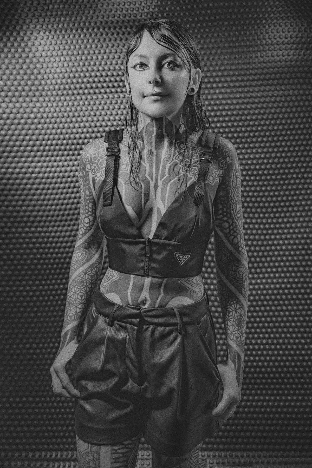 Black and white portrait of Krista in a cropped top and shorts against a textured metal wall