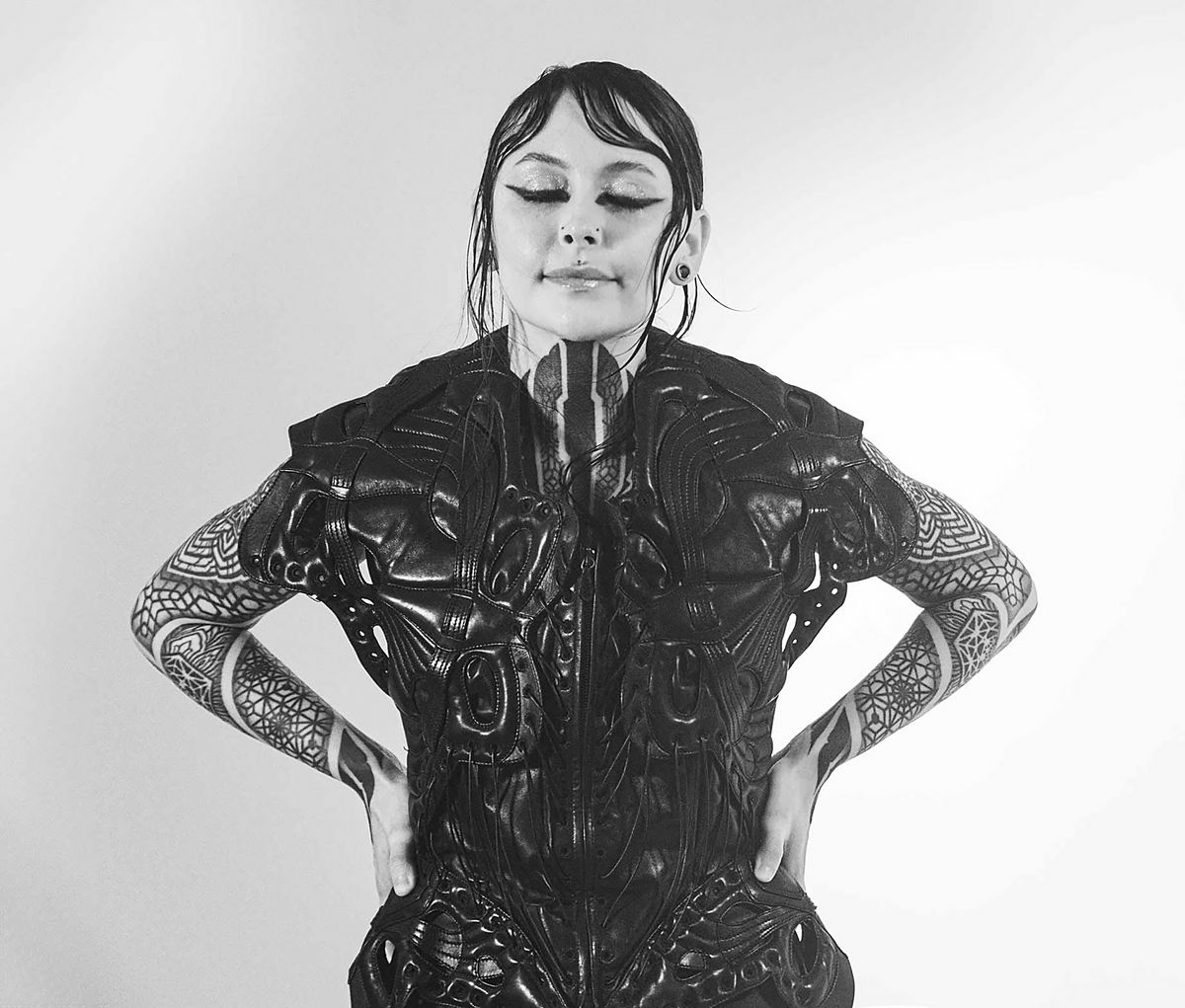 Black and white portrait of Krista with eyes closed, wearing a sculptural top with her hands on her hips