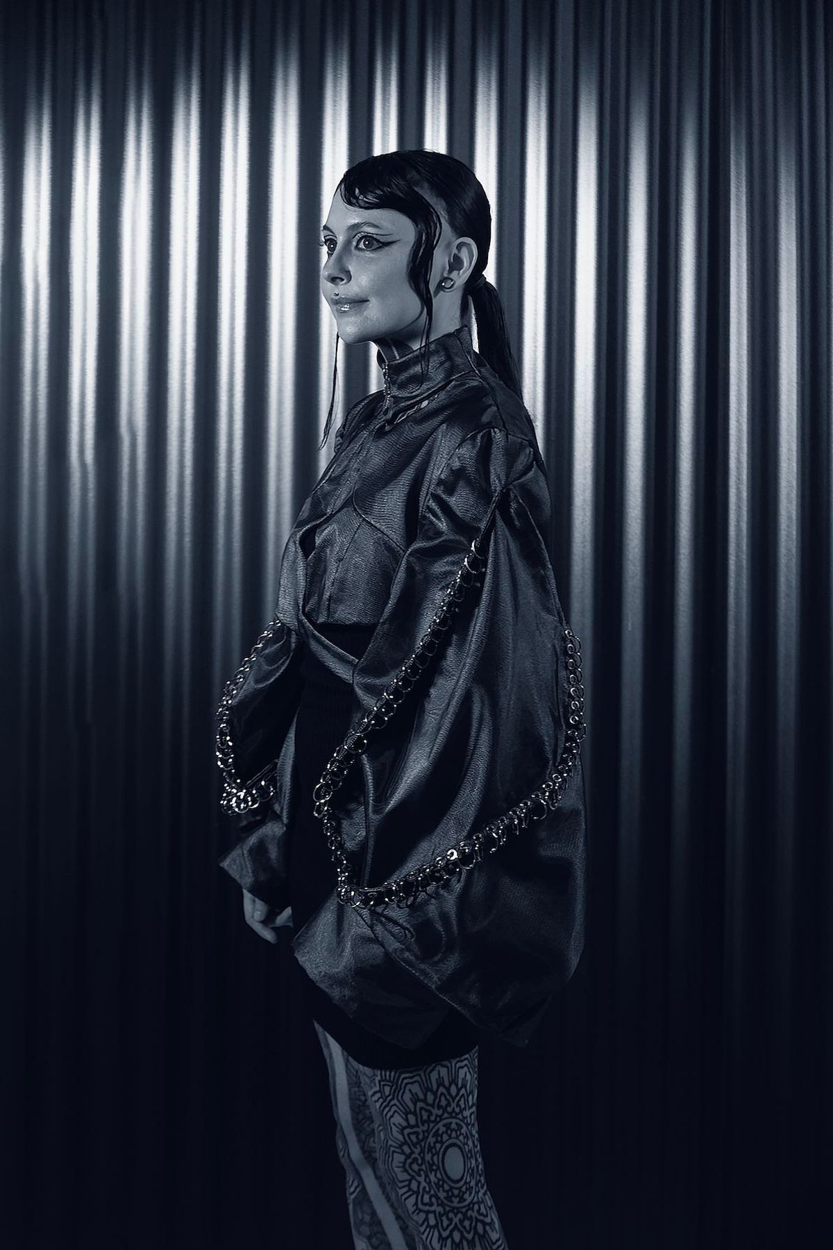 Krista in an oversized dark jacket, standing sideways against a corrugated metal backdrop
