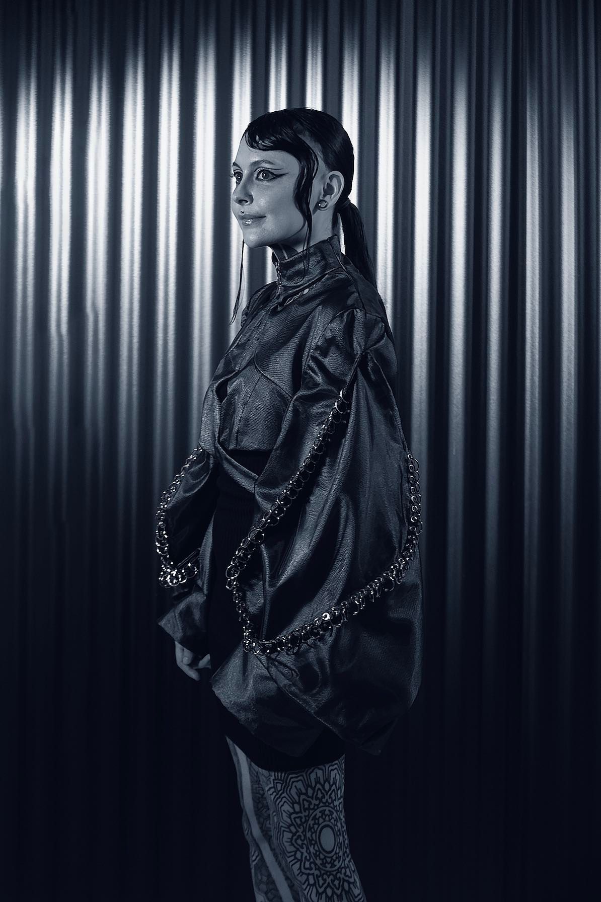 Krista in an oversized dark jacket, standing sideways against a corrugated metal backdrop