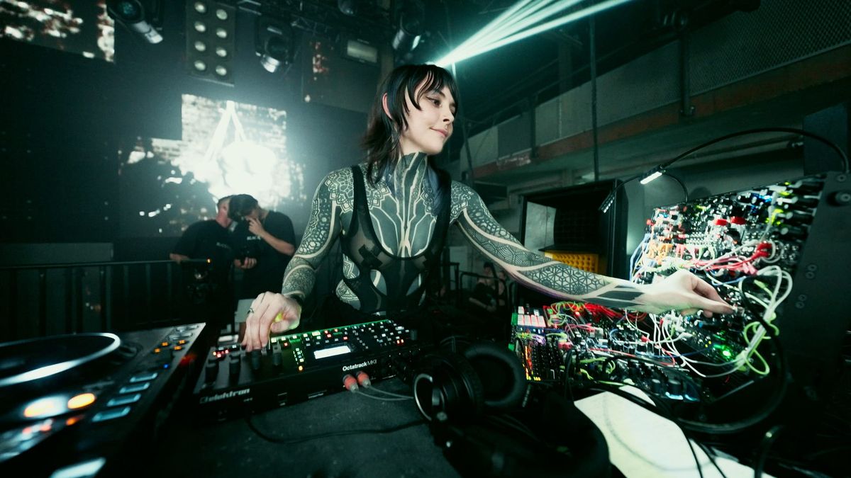 Krista performing live at a modular synthesizer and mixer on a dark club stage, with laser light and stage crew behind her