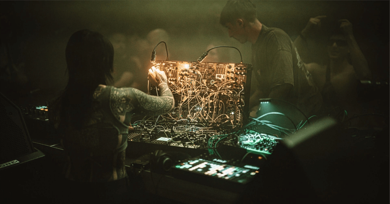Tattooed hand adjusting cables on a modular synthesizer at a hazy live performance, with a second performer beside them