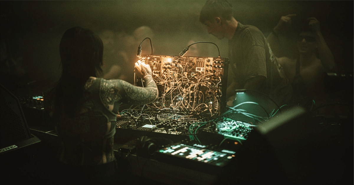 Tattooed hand adjusting cables on a modular synthesizer at a hazy live performance, with a second performer beside them