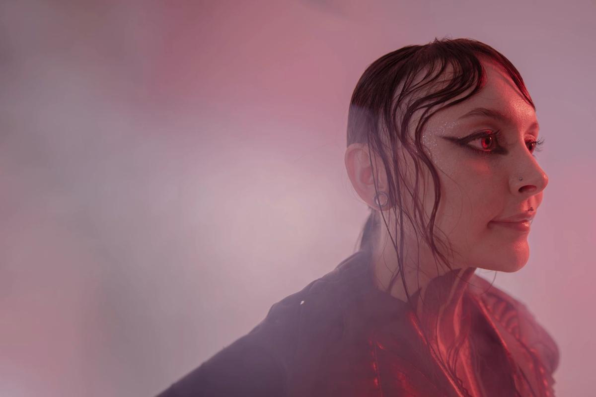 Krista in profile with a raised collar, lit by a soft red glow through studio haze