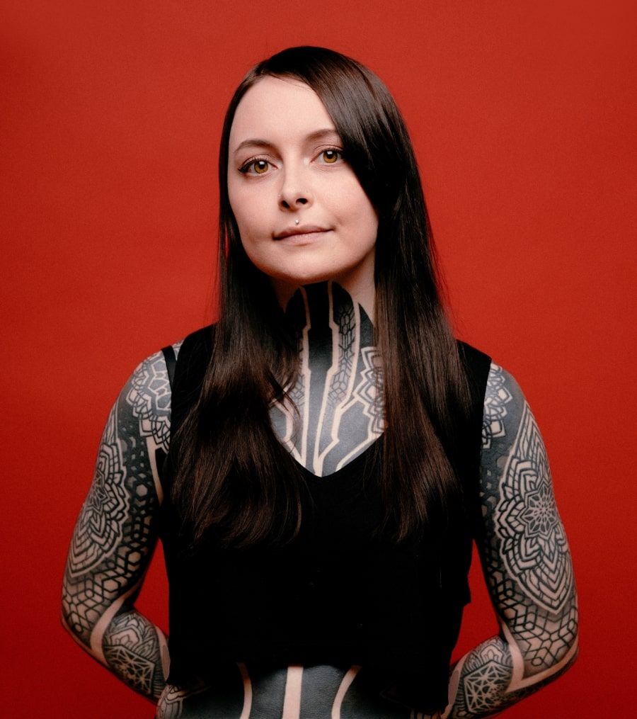 Krista with long dark hair and tattooed arms, wearing a black sleeveless top against a red wall