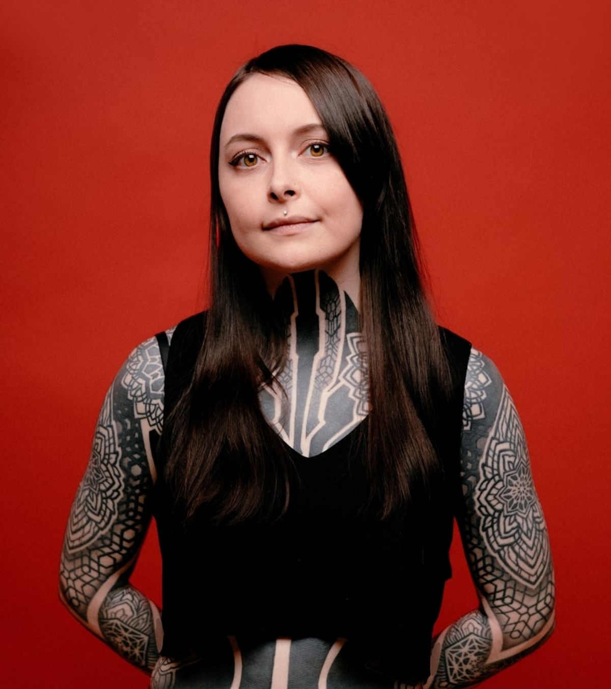 Krista with long dark hair and tattooed arms, wearing a black sleeveless top against a red wall