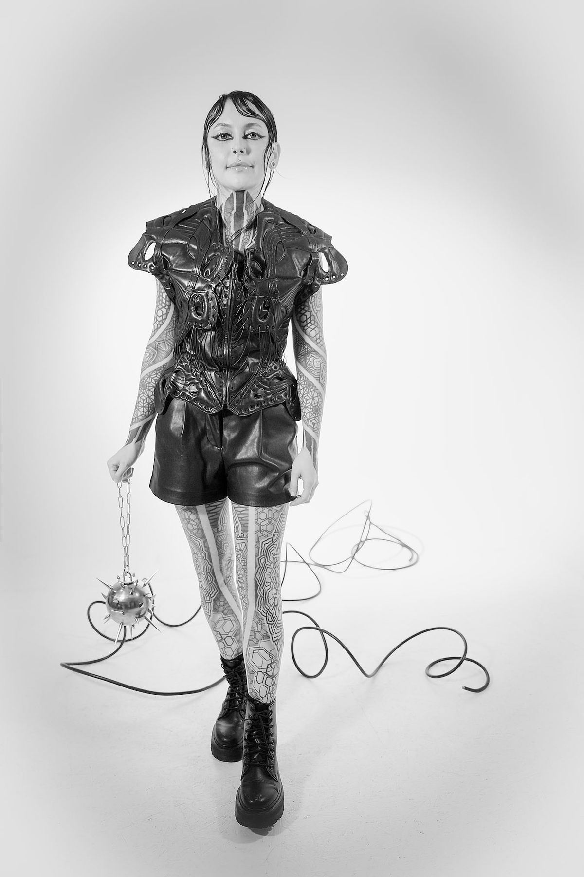 Full-length black and white portrait of Krista in a sculptural top, shorts and boots, holding trailing cables
