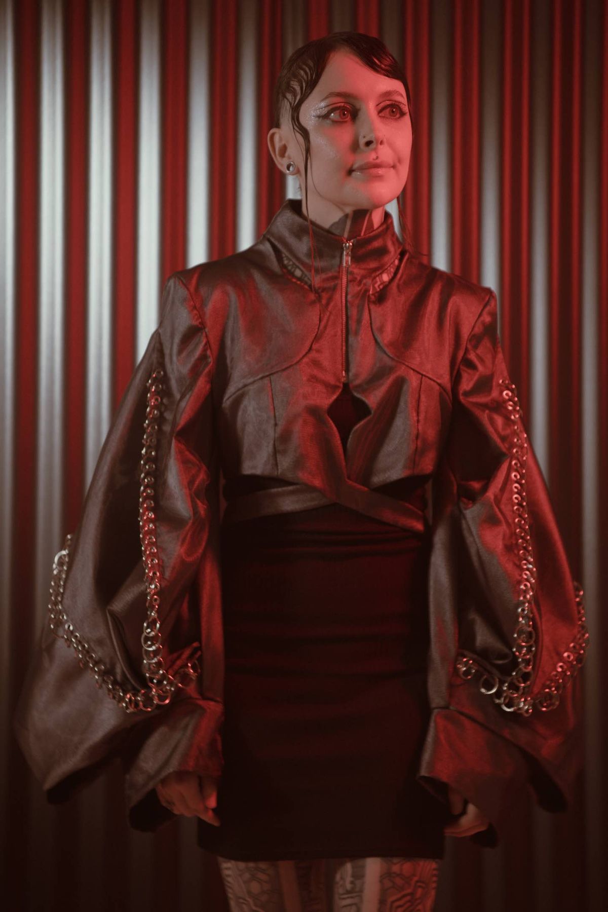 Krista standing in a metallic jacket and dark skirt against a corrugated backdrop under red light