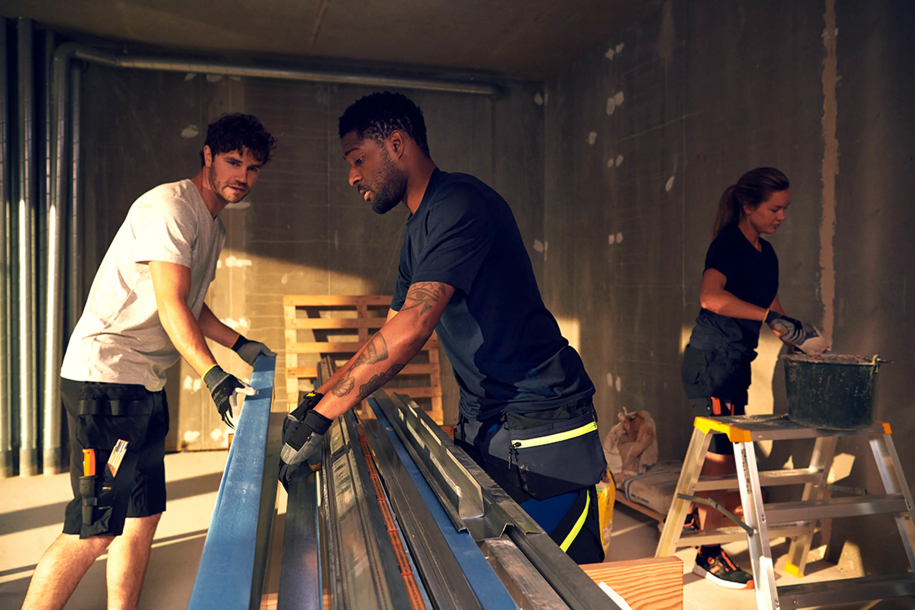 Tre håndverkere i shorts jobber innendørs på en byggeplass