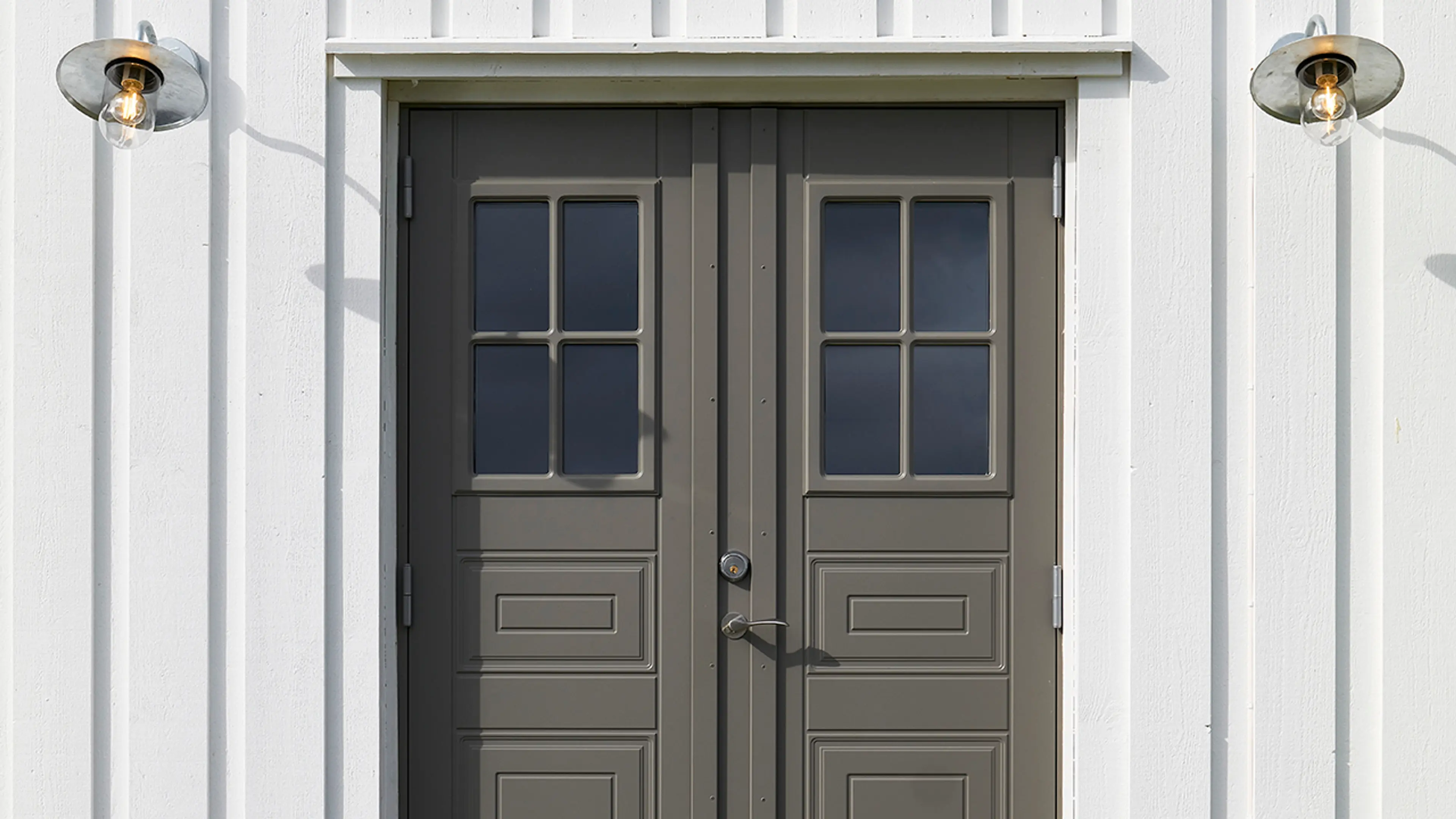 Stående kledning som er hvitmalt med profilerte dobbeltdører og to lamper ved inngangen.