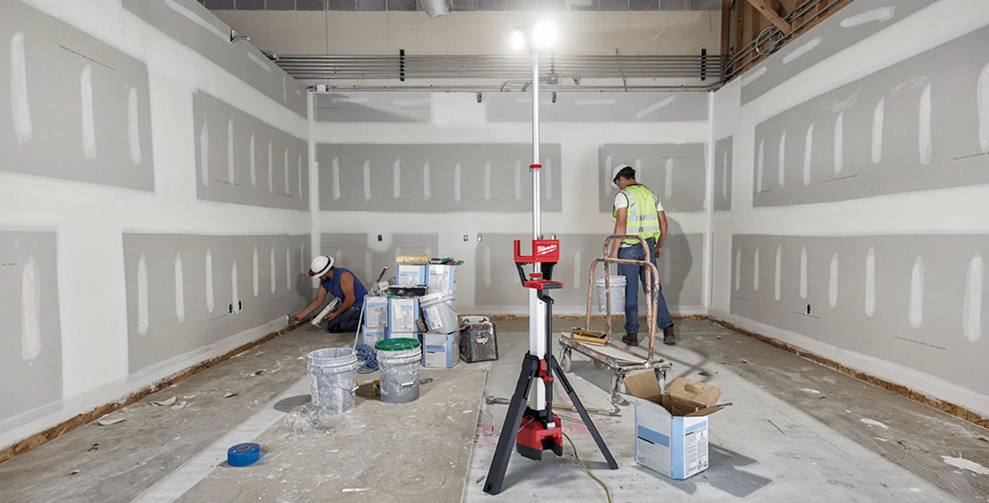 To håndverkere i rom som bygges og M18 HOSALC arbeidslampe fra MILWAUKEE® som lyser opp.