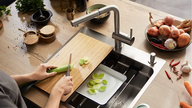 Person som kutter grønnsaker på skjærebrett i kjøkkenvasken med rist og oppsamlingsskål.