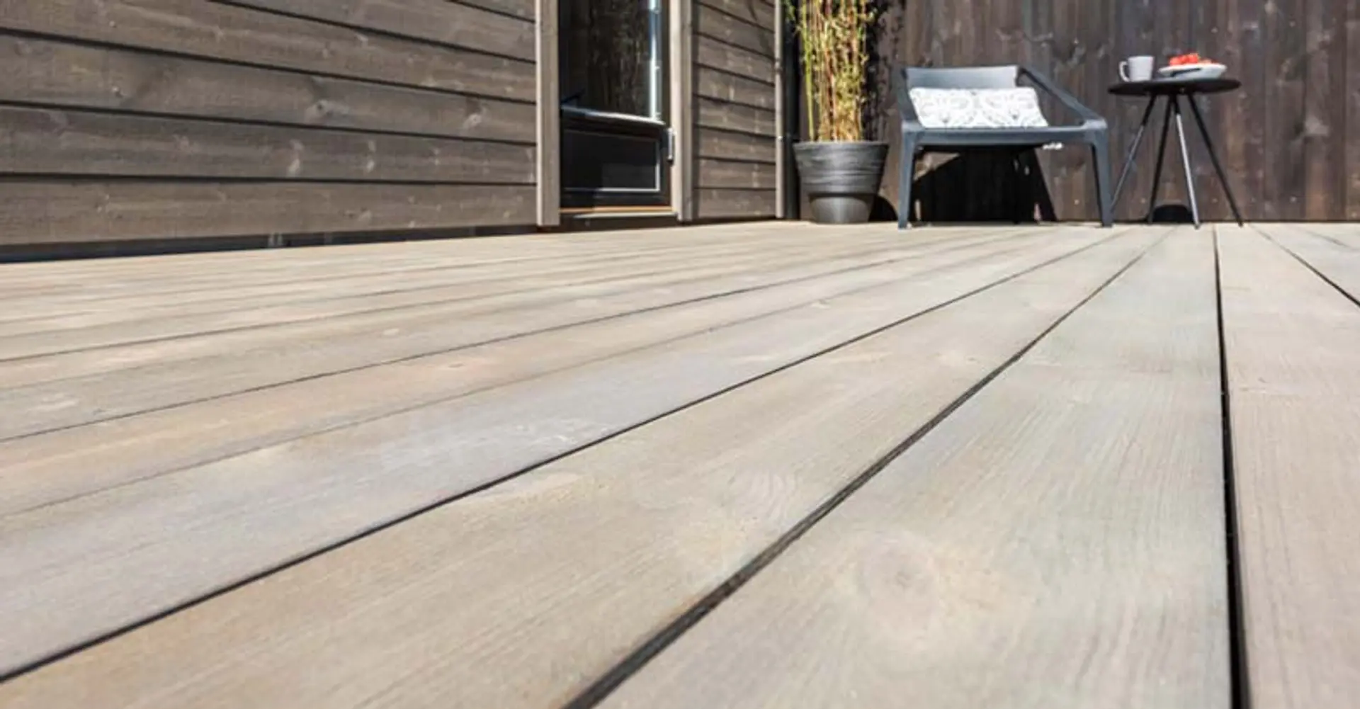 Nærbilde av grå terrassebord med hytta og hagemøbler i bakgrunn.