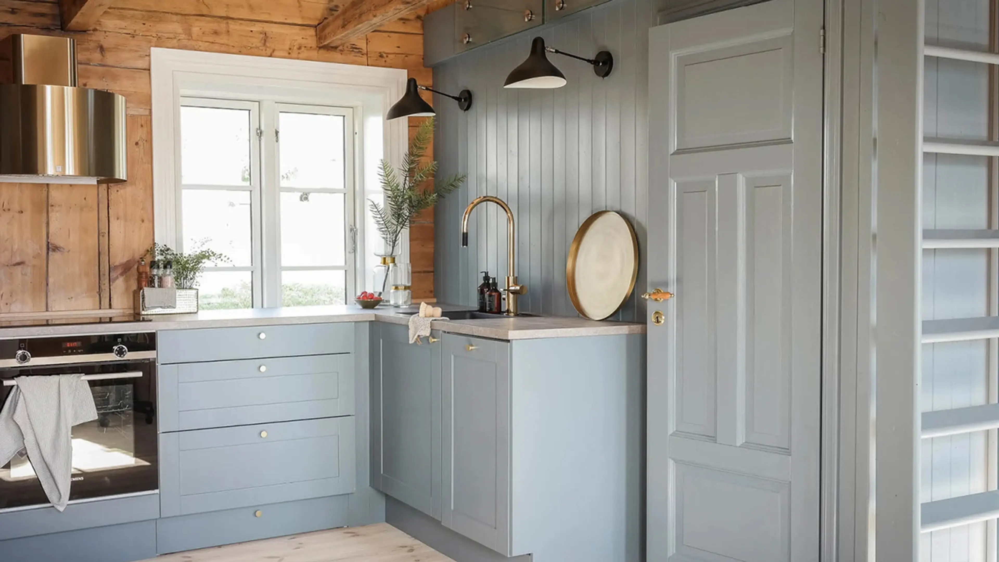 Hyttekjøkken med kjøkkenhette i messing, innebygd stekeovn og fronter malt i fargen 5044 Isblå fra Jotun.
