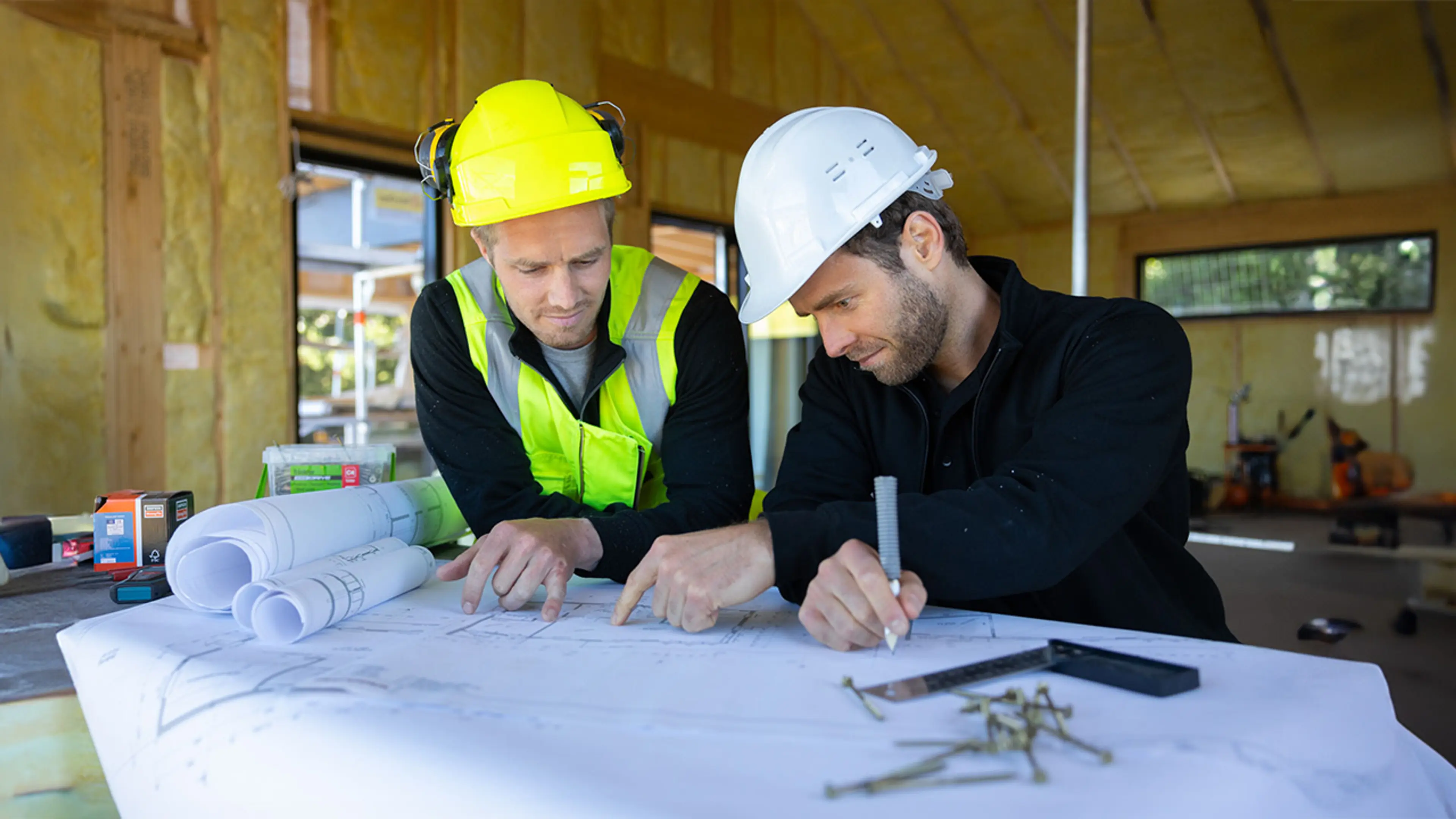 To menn som ser på plantegninger inne i et hus under oppbygging.