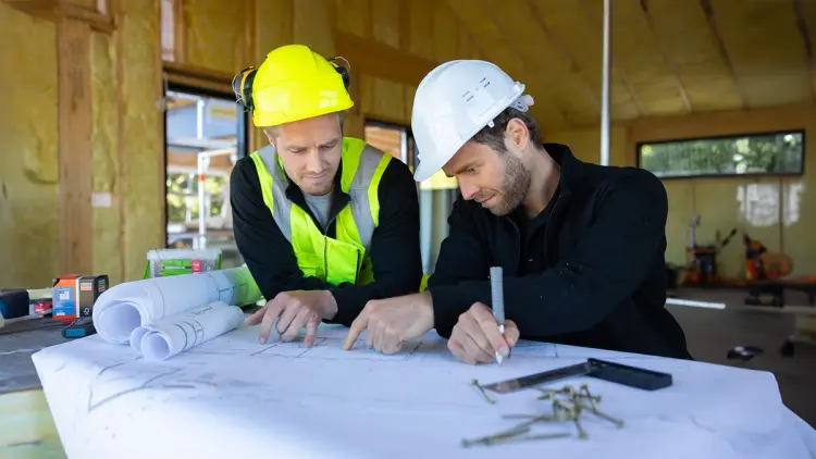 To menn som ser på plantegninger inne i et hus under oppbygging.