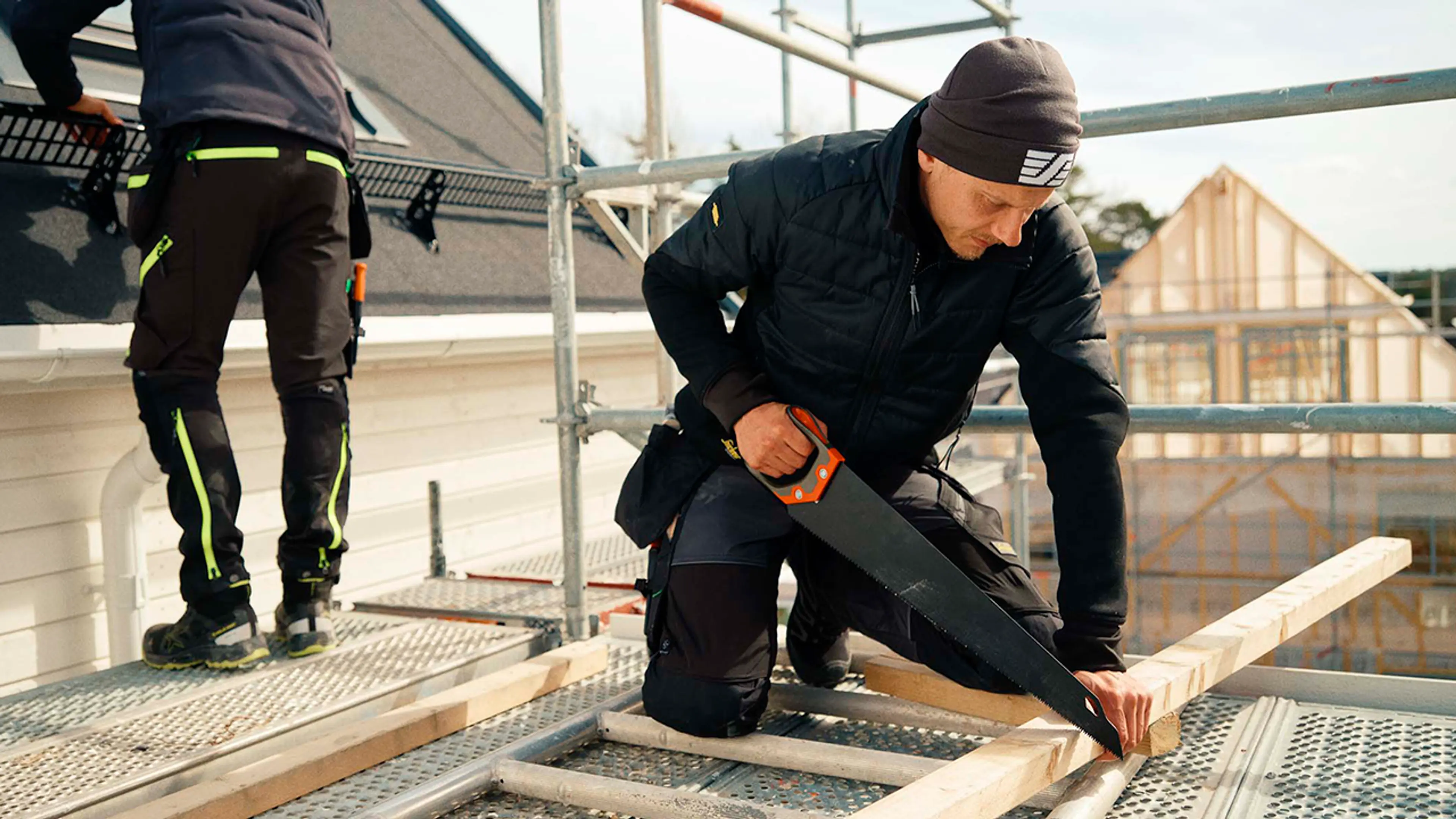 Håndverkere som arbeider med tak og sager på stillas med arbeidsklær.
