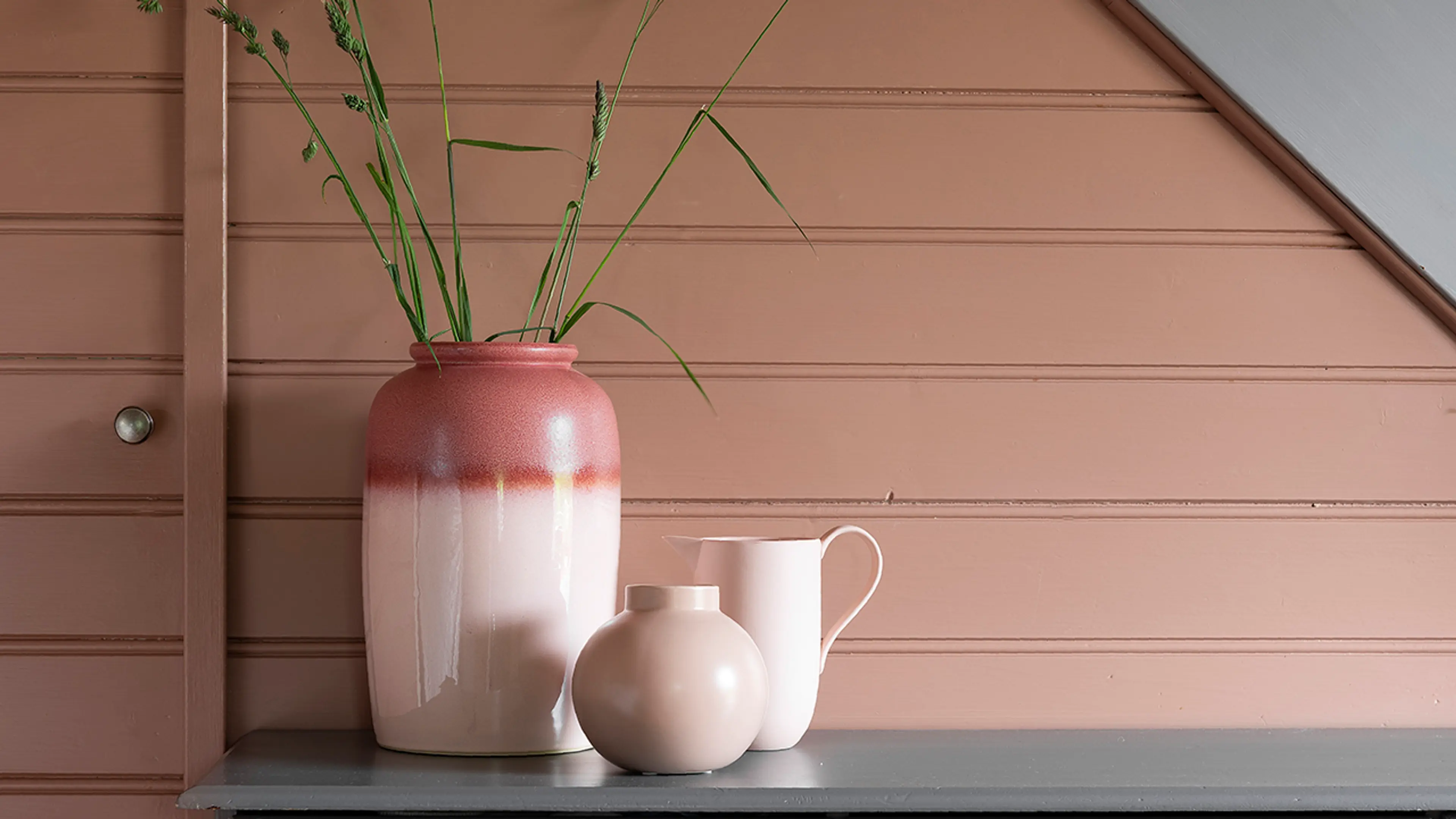 Nærbilde av liggende trepanel som er malt i en dus rosafarge. Foran står en gråmalt kommode med vaser og blomster på.