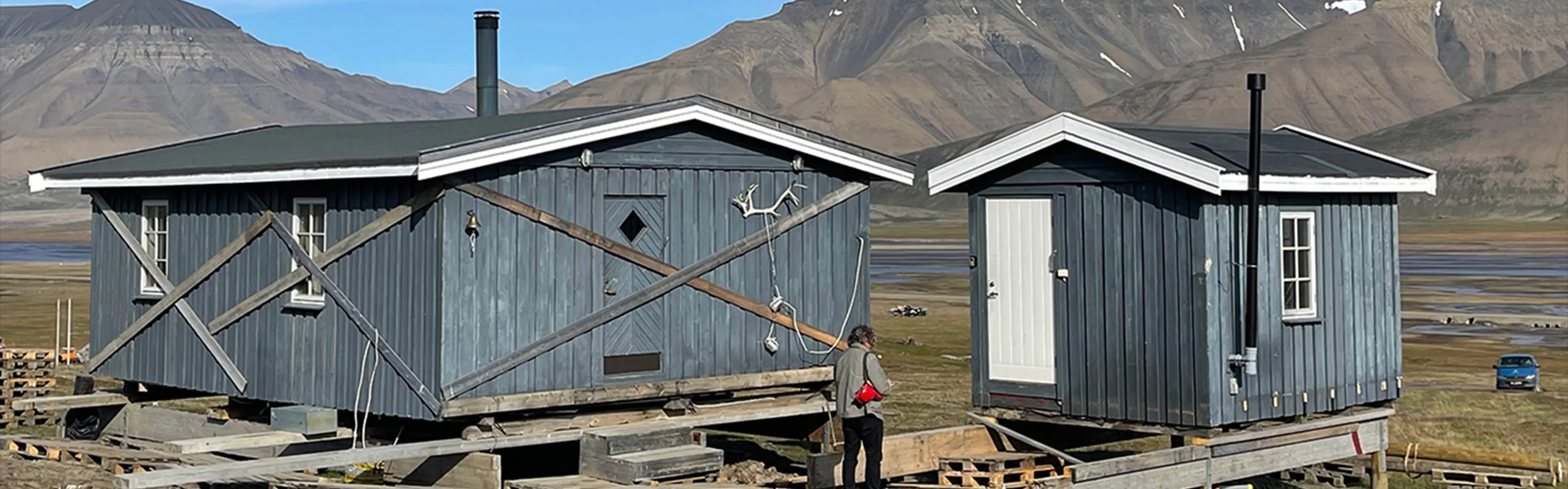 To blå hytter i naturen med en mann som står foran og fjell i bakgrunn.