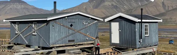 To blå hytter i naturen med en mann som står foran og fjell i bakgrunn.
