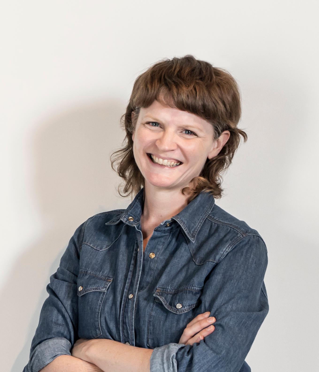 Headshot of visual artist Caitlin Franzmann. Caitlin is wearing a denim jacket with arms folded and smiling broadly at the camera. She stands in front of a white background.