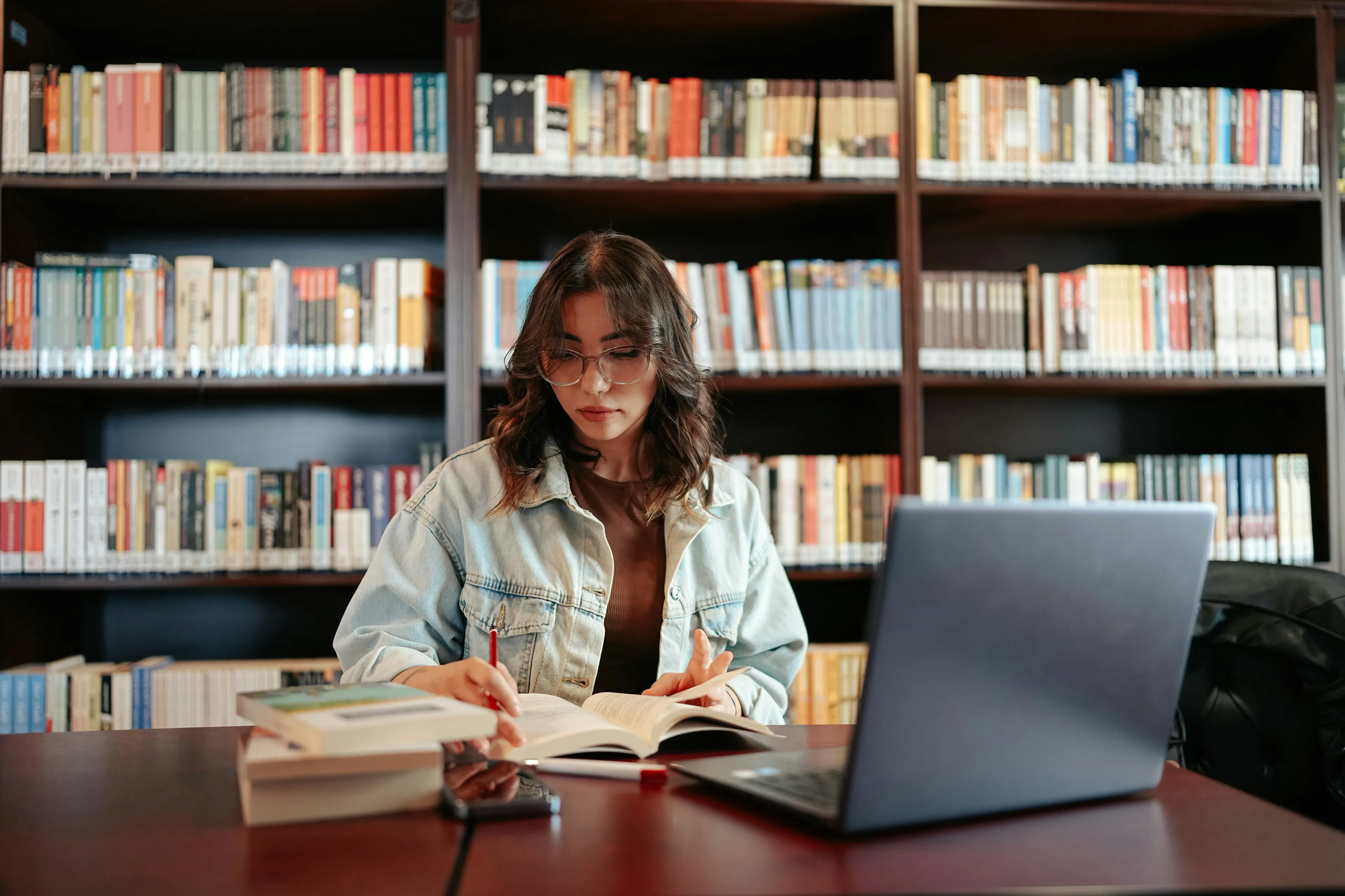 Ung kvinne med briller studerer i bibliotek, omgitt av bøker og med en bærbar PC foran seg. Hun skriver i en bok og ser fokusert ut.
