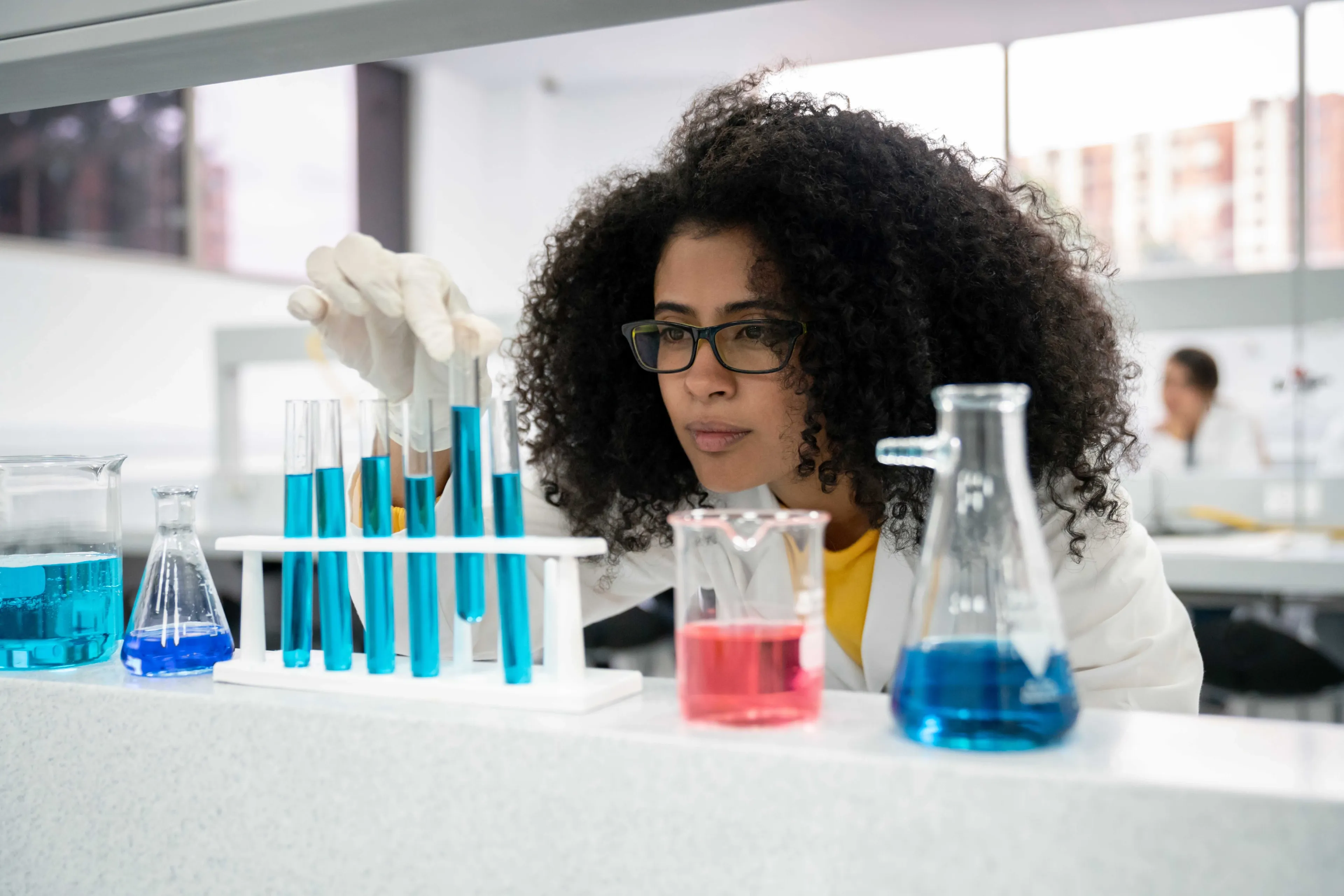 Kjemistudent med briller og laboratoriefrakk arbeider konsentrert i et laboratorium. Hun holder et reagensrør med blå væske og studerer det nøye.