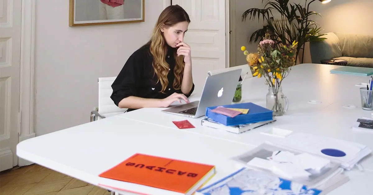 Kvinne jobber fokusert på en laptop ved et ryddig bord med bøker, papirer og en blomsterbukett.