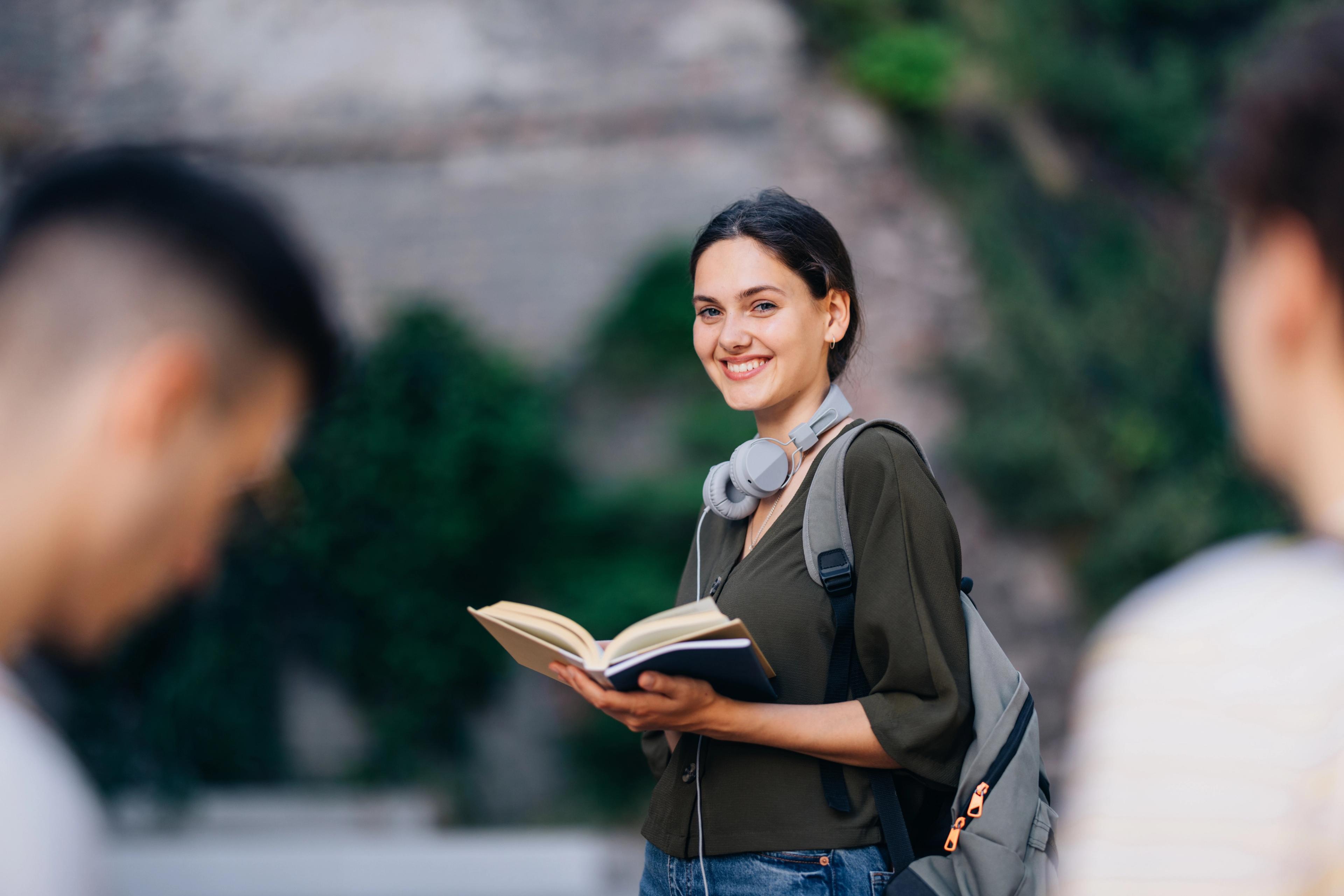 Smilende Sonans-student med ryggsekk, holder en åpen bok utendørs, med andre studenter i bakgrunnen.