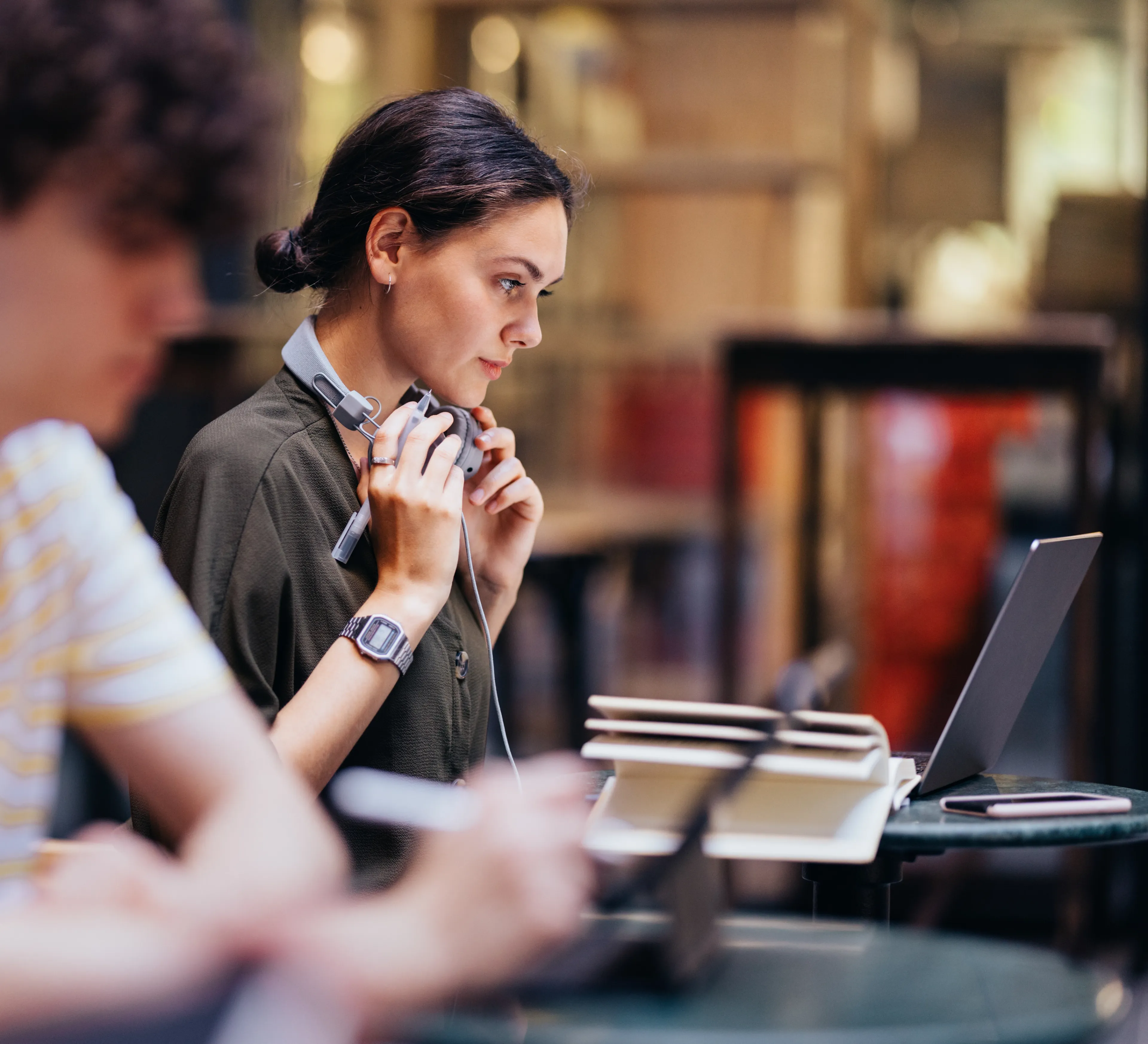 Student med hodetelefoner rundt halsen studerer på nett med laptop og bøker på lesesalen.