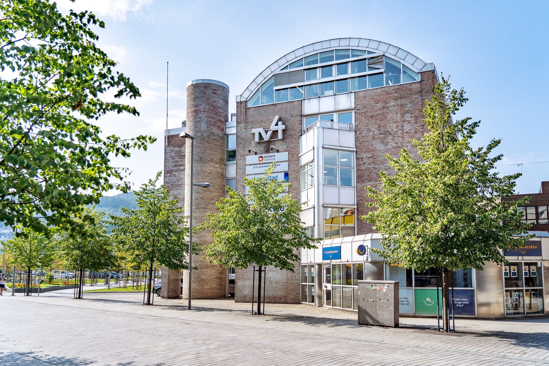 Bygning i Drammen sentrum med Sonans-skilt på fasaden, omgitt av grønne trær under blå himmel.