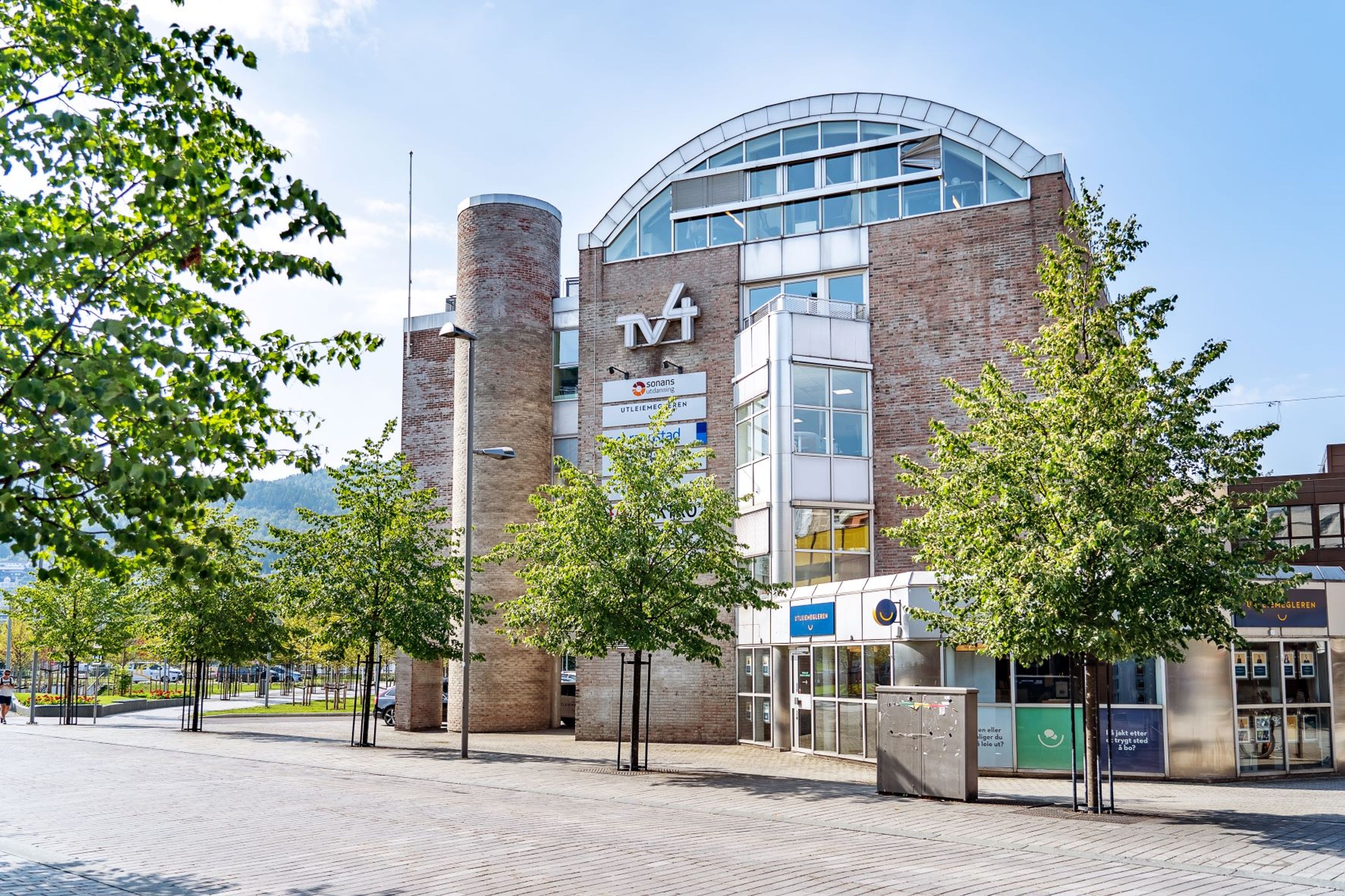 Bygning i Drammen sentrum med Sonans-skilt på fasaden, omgitt av grønne trær under blå himmel.