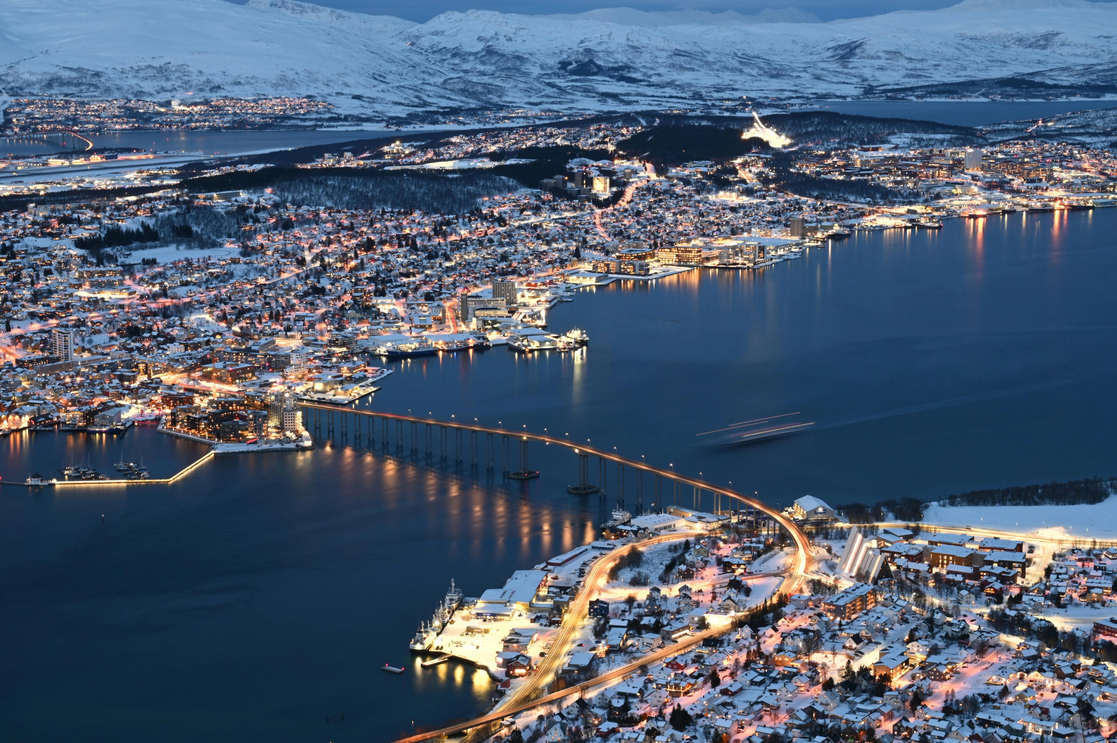 Tromsø by om vinteren, med lys over byen, broen og snødekte fjell i bakgrunnen.