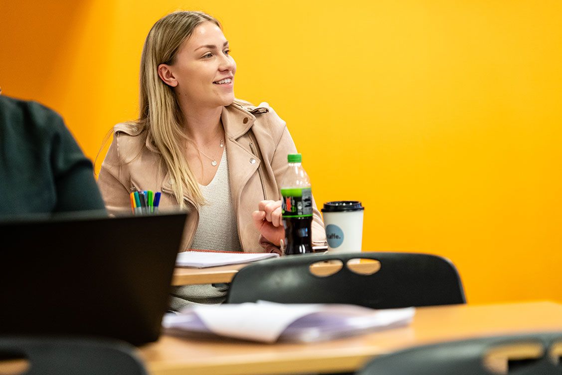 Smilende student i klasserom med notater, penner og drikke foran seg, mot en gul vegg.