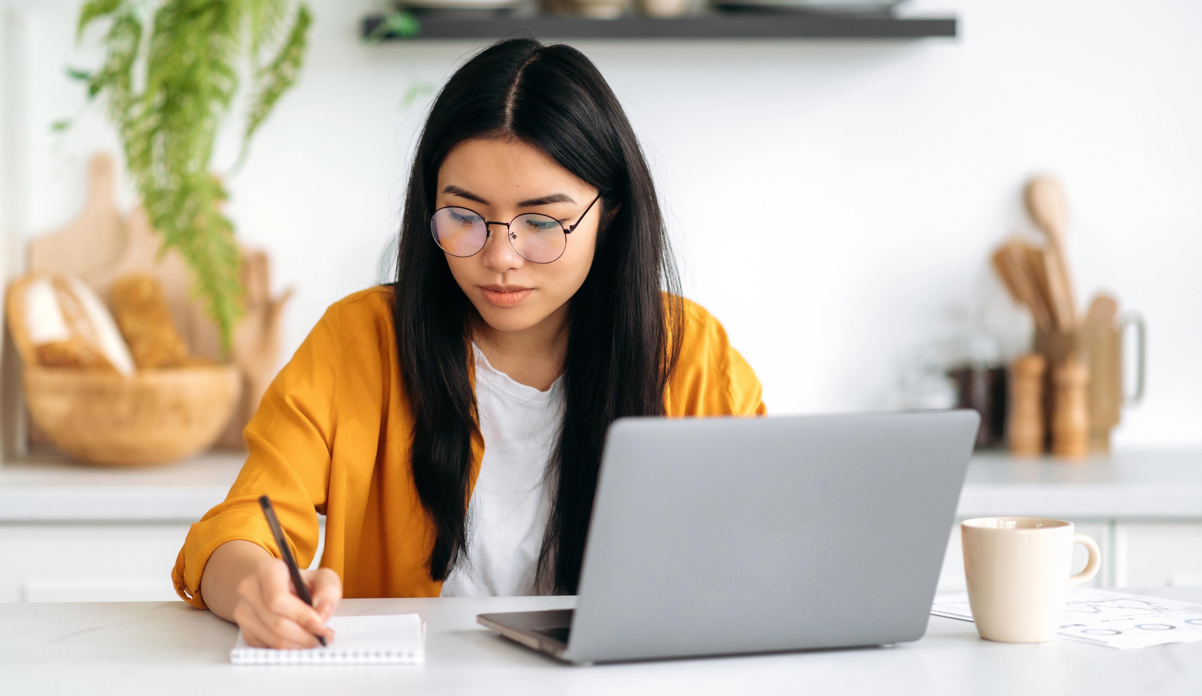 kvinnelig student sitter konsentrert foran en pc og noterer på en skriveblokk.
