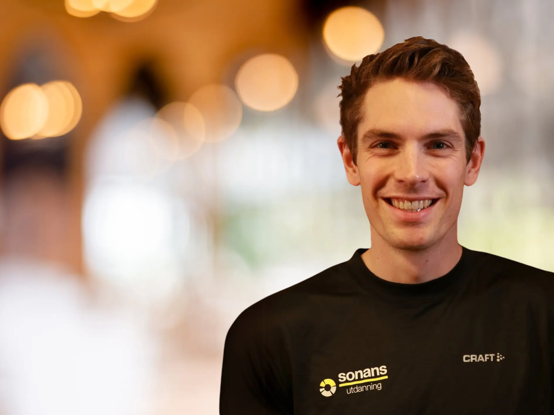 Smilende mann i svart Sonans-treningsskjorte med uskarp bakgrunn og varmhvite lys.