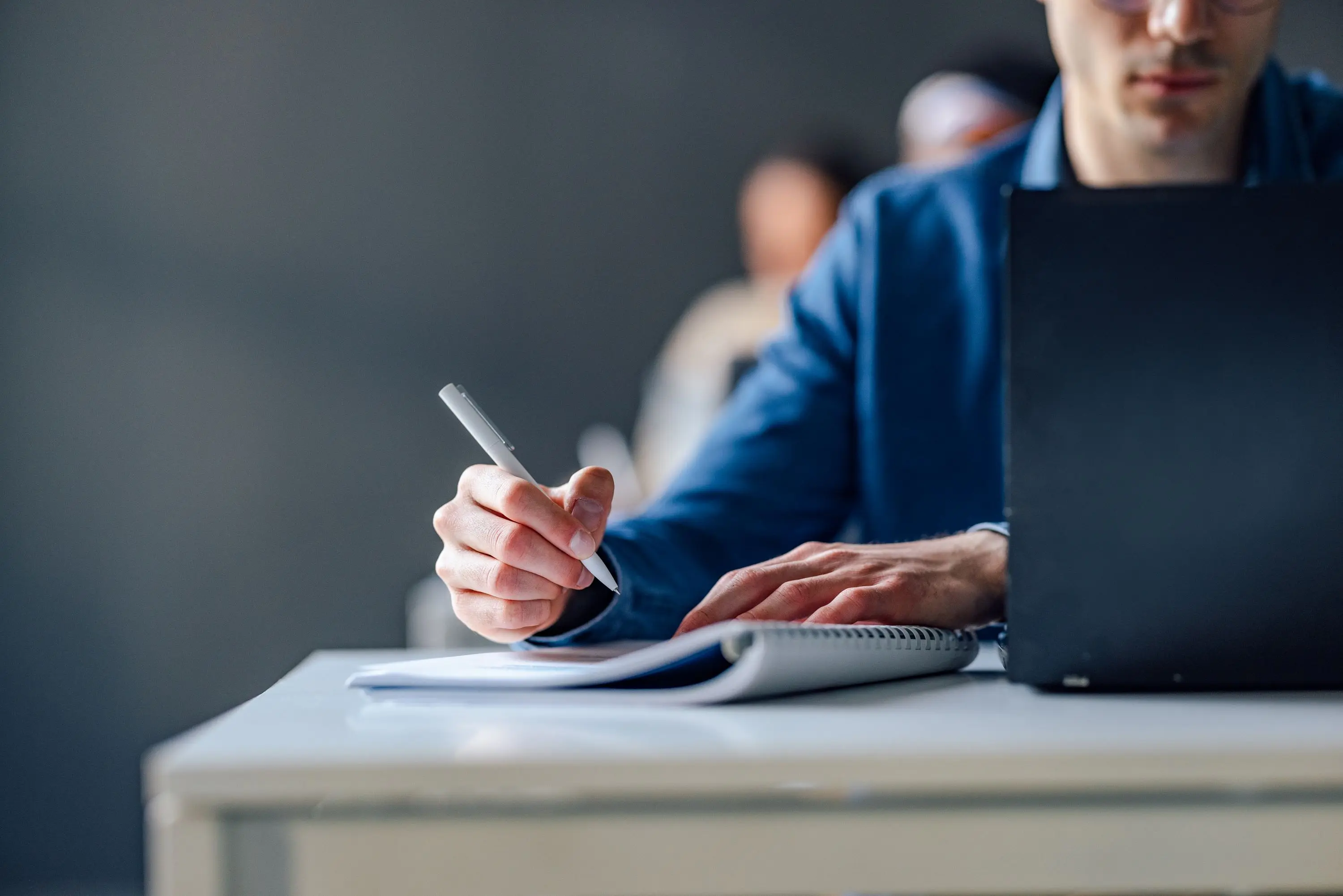 Student i blå skjorte tar notater i en spiralblokk mens han jobber på en bærbar PC i et klasserom.