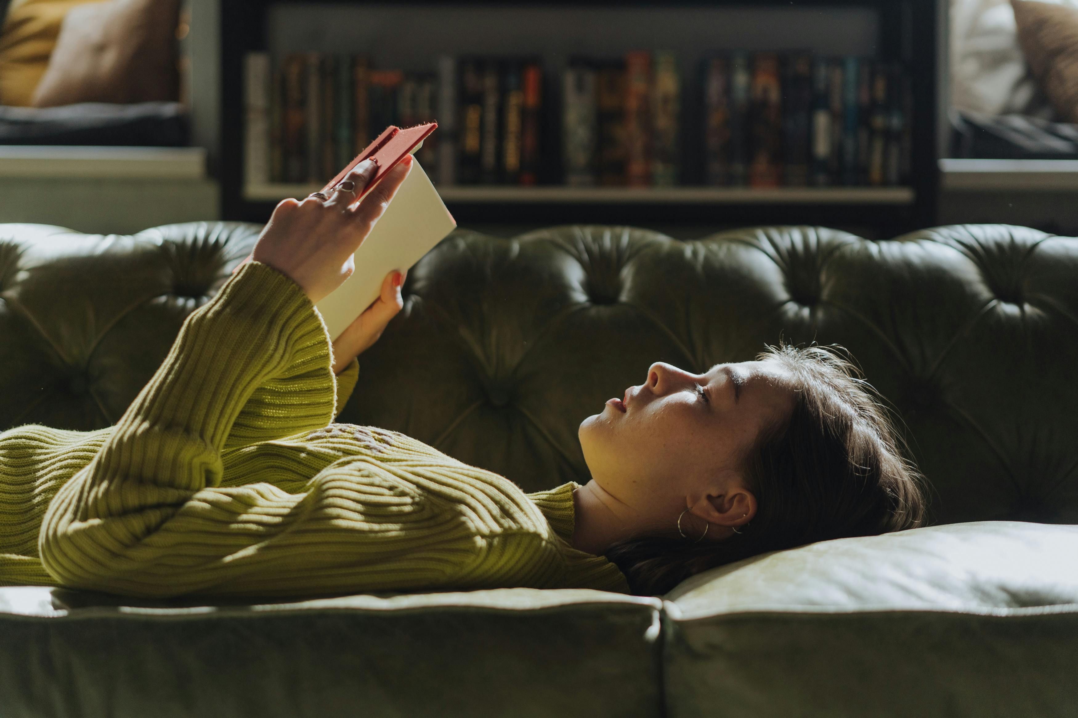 Student som ligger på en grønn sofa, leser en bok og studerer til eksamen.