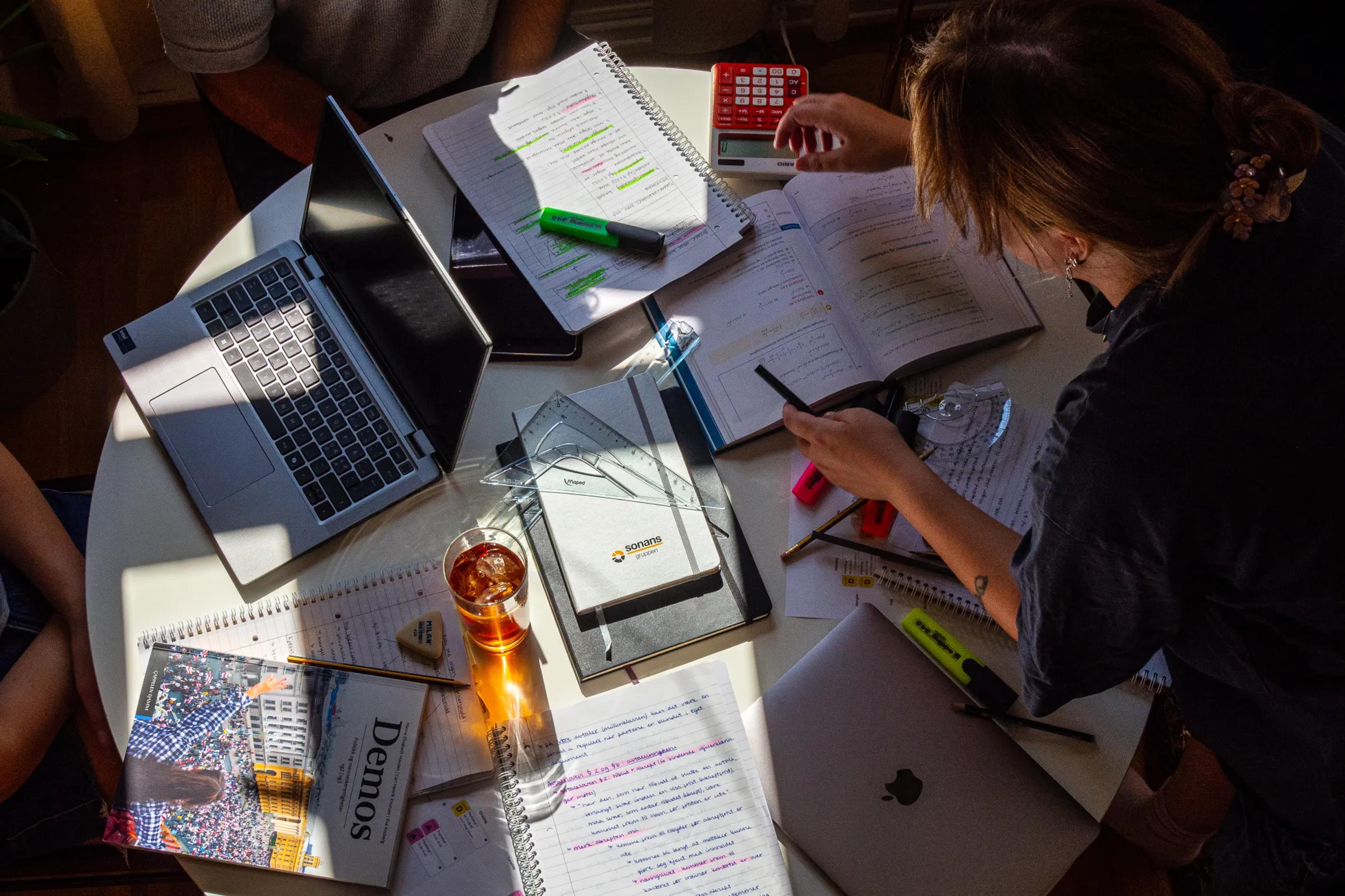 Studenter jobber sammen rundt et bord fullt av skolebøker, markeringstusjer, notater og digitale hjelpemidler - med Sonans-blokk synlig i midten.