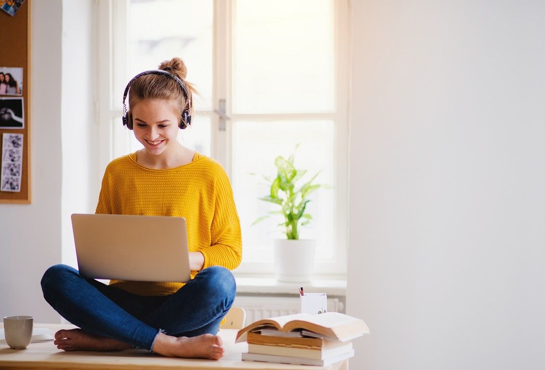 Smilende kvinne i gult sitter med laptop og hodetelefoner, omgitt av bøker, i et lyst rom.
