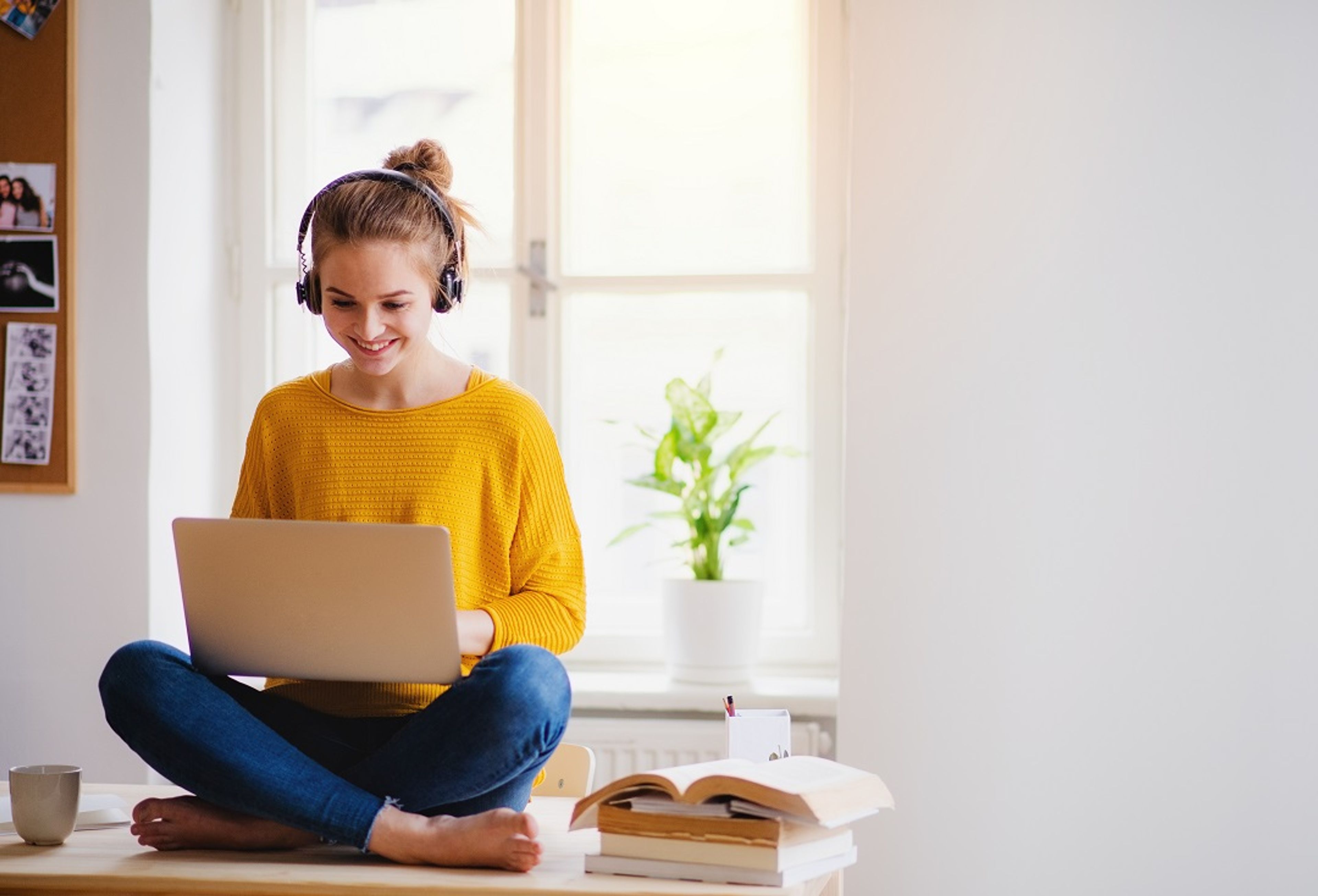 Smilende kvinne i gult sitter med laptop og hodetelefoner, omgitt av bøker, i et lyst rom.