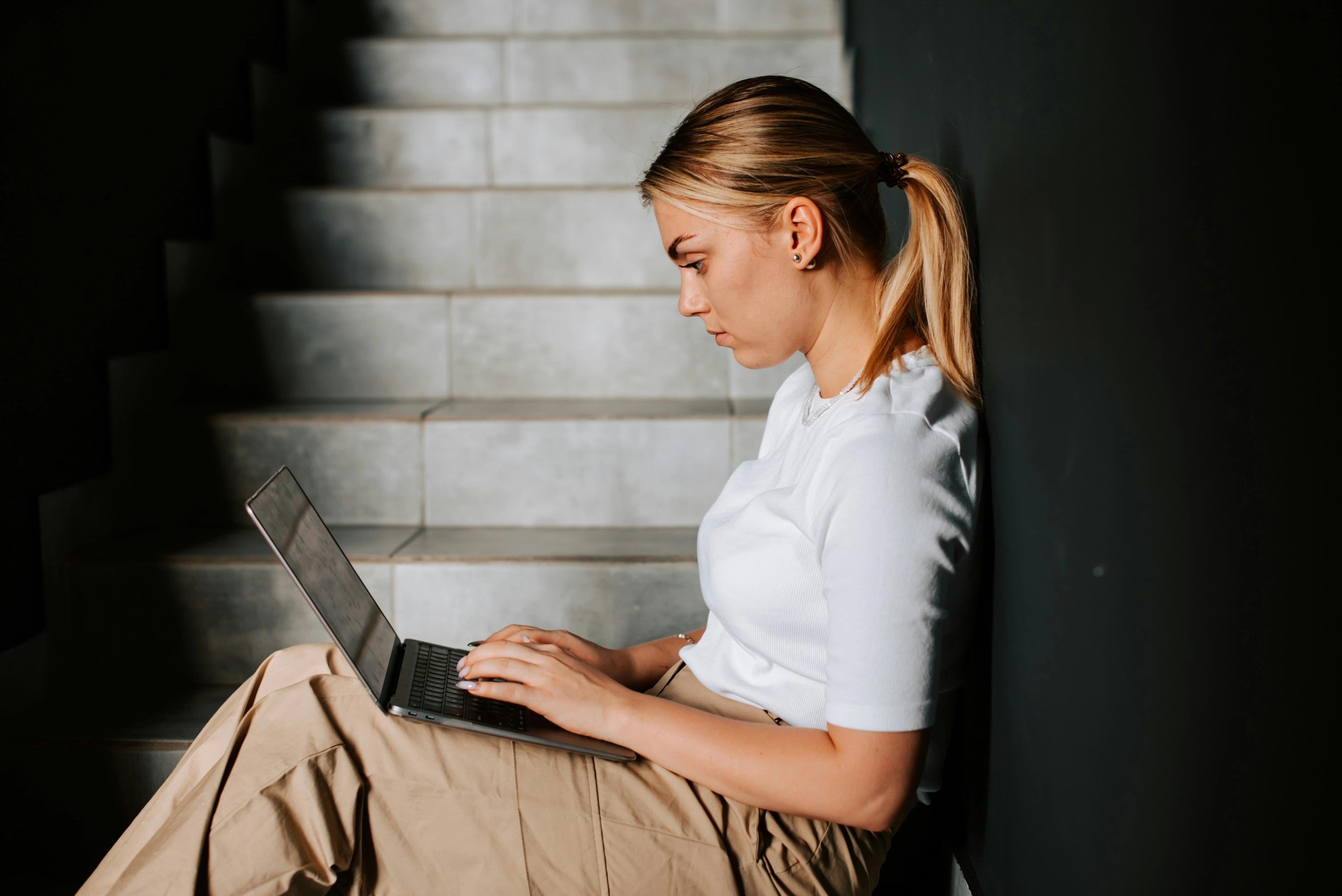Ung kvinne i hvit topp og beige bukse jobber på en laptop mens hun sitter i en trapp.