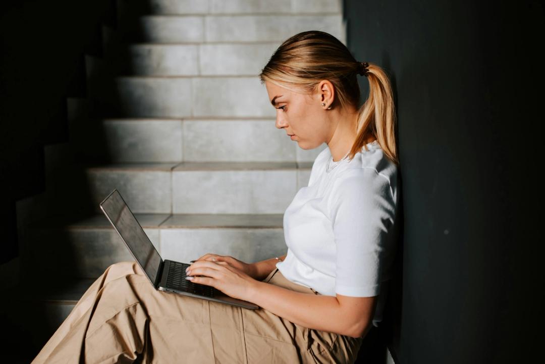 Ung kvinne i hvit topp og beige bukse jobber på en laptop mens hun sitter i en trapp.