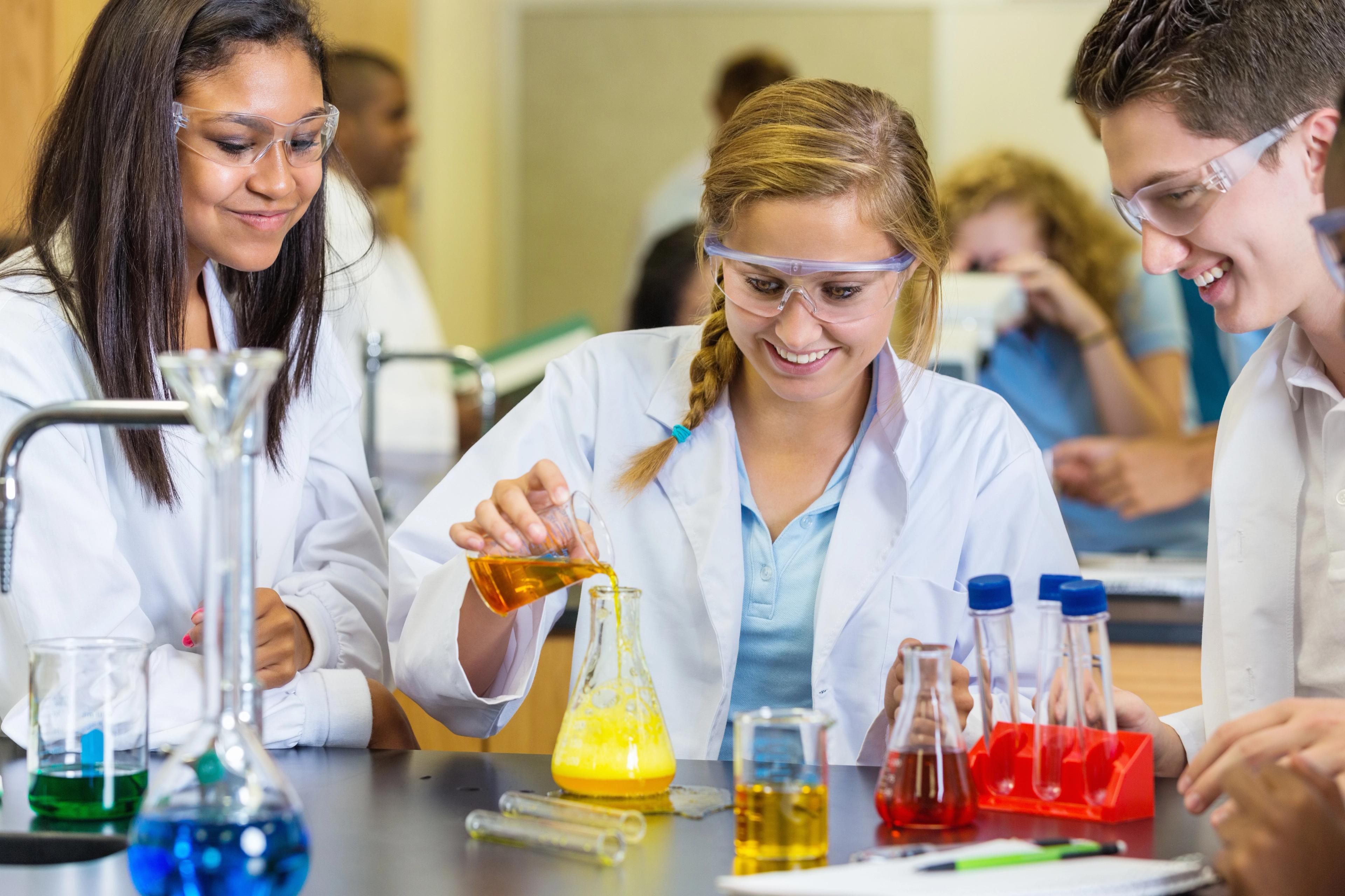 Student i hvit labfrakk og vernebriller utfører kjemisk forsøk med reagensrør fylt med fargede væsker i laboratorium.