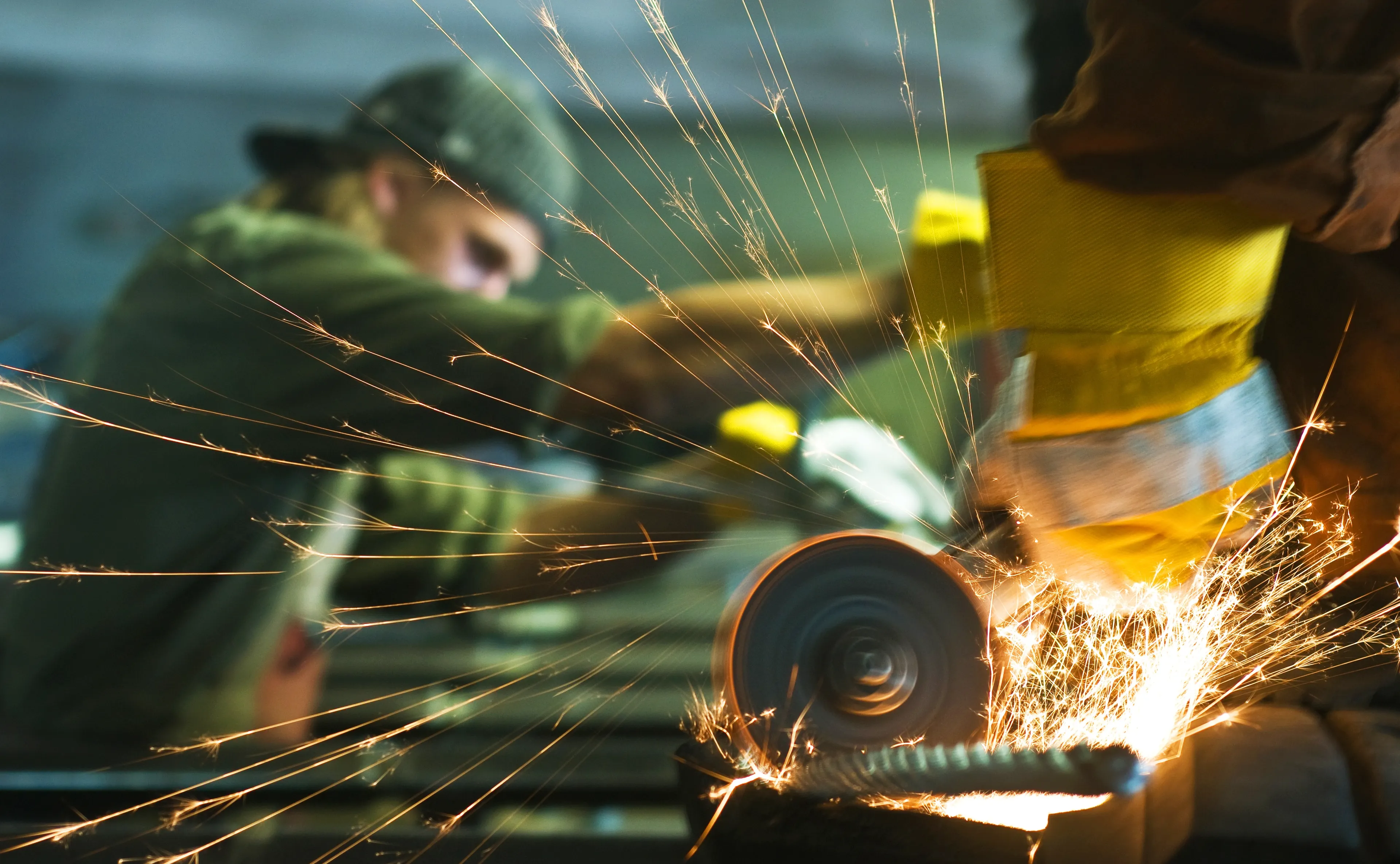To personer arbeider med metall i et verksted. Den ene bruker en vinkelsliper, som skaper gnister, mens den andre holder i arbeidsstykket.