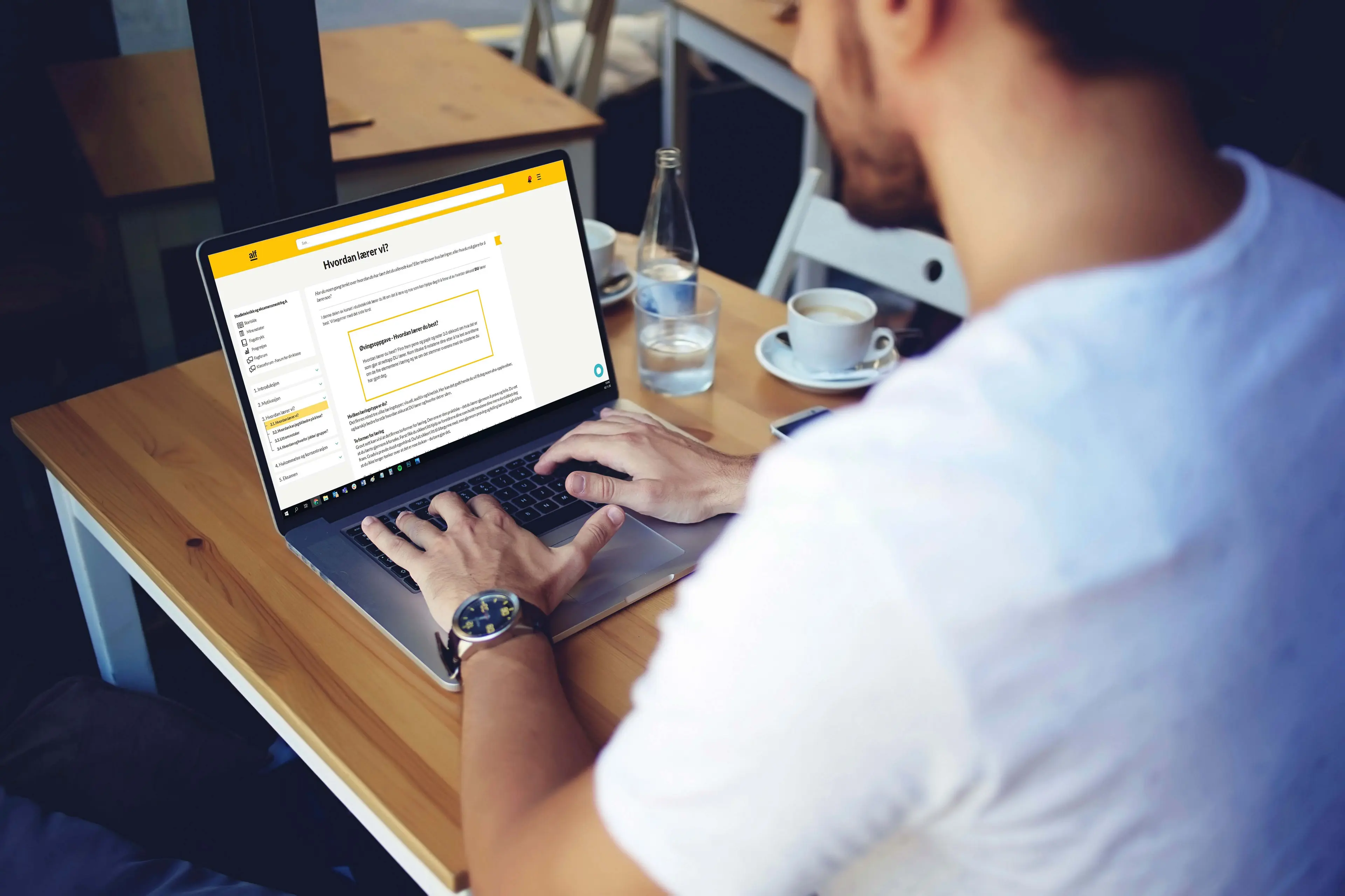 Mann i hvit t-skjorte studerer på laptop med kaffe ved et kafébord, mens Sonans sin læreringsplattform "ALF" er åpen.