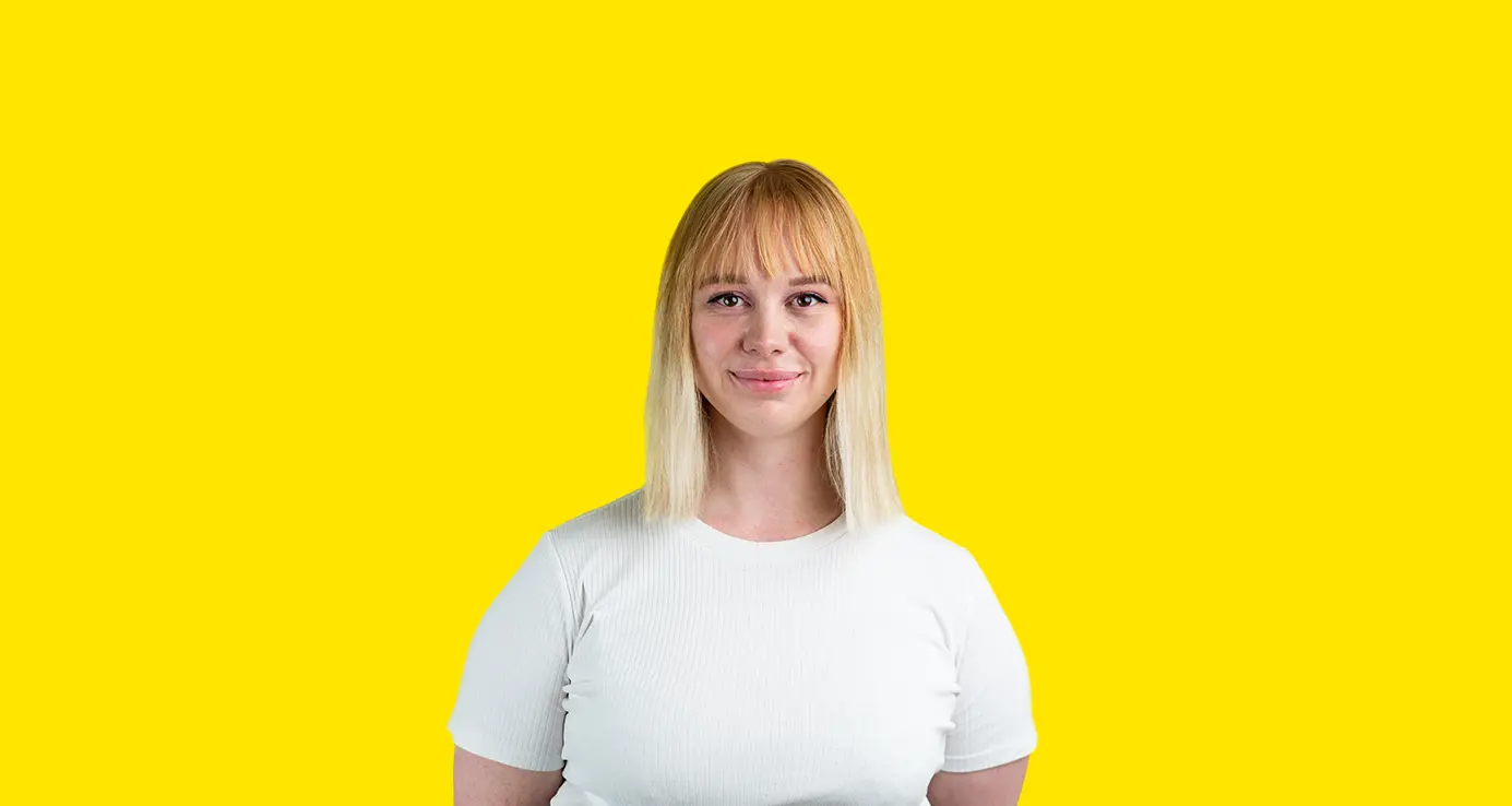 Ung kvinne med blondt hår og hvit t-skjorte smiler foran en gul bakgrunn.