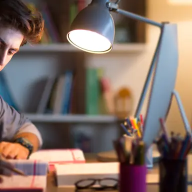 Student som sitter ved et skrivebord og studerer. Han er omgitt av bøker, en dataskjerm, skrivebordslampe og penneholdere.