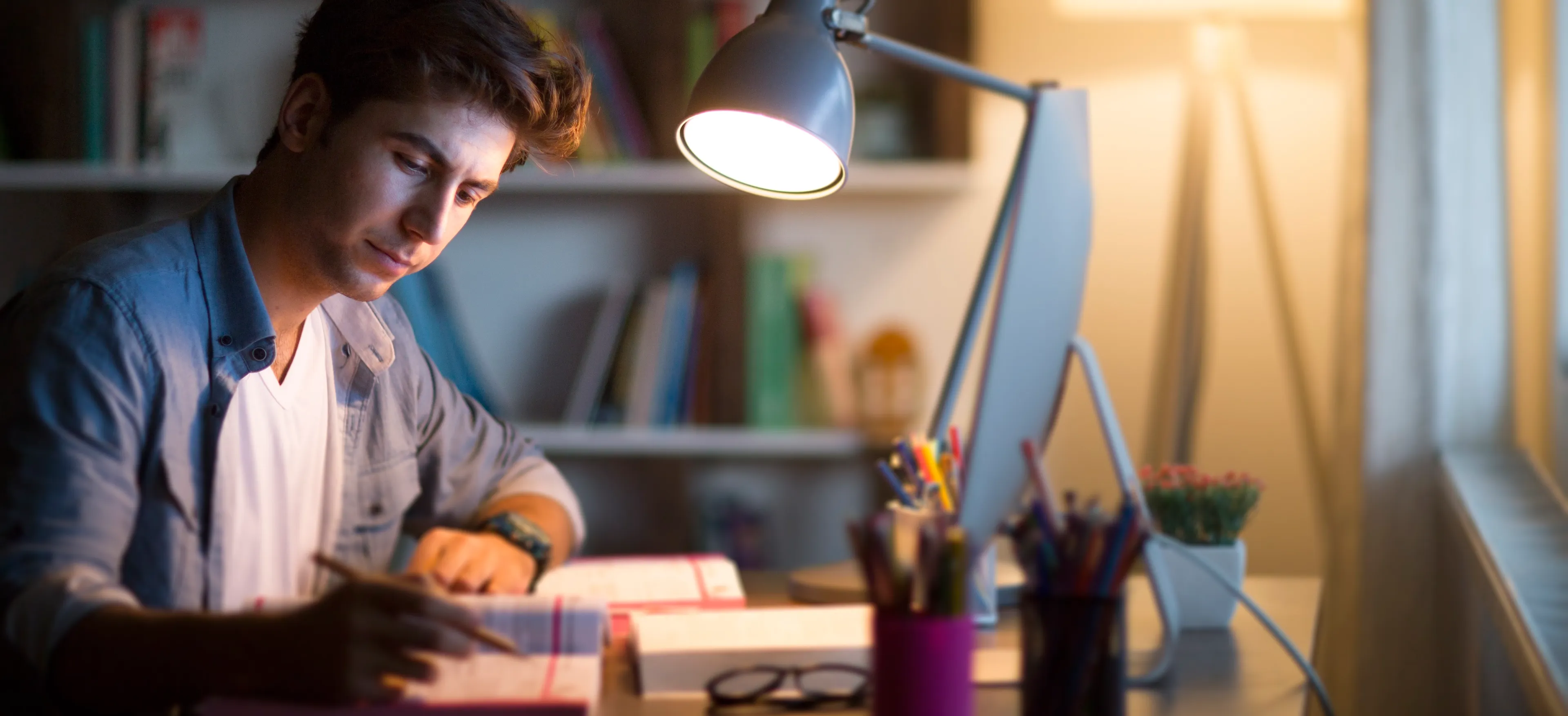 Student som sitter ved et skrivebord og studerer. Han er omgitt av bøker, en dataskjerm, skrivebordslampe og penneholdere.